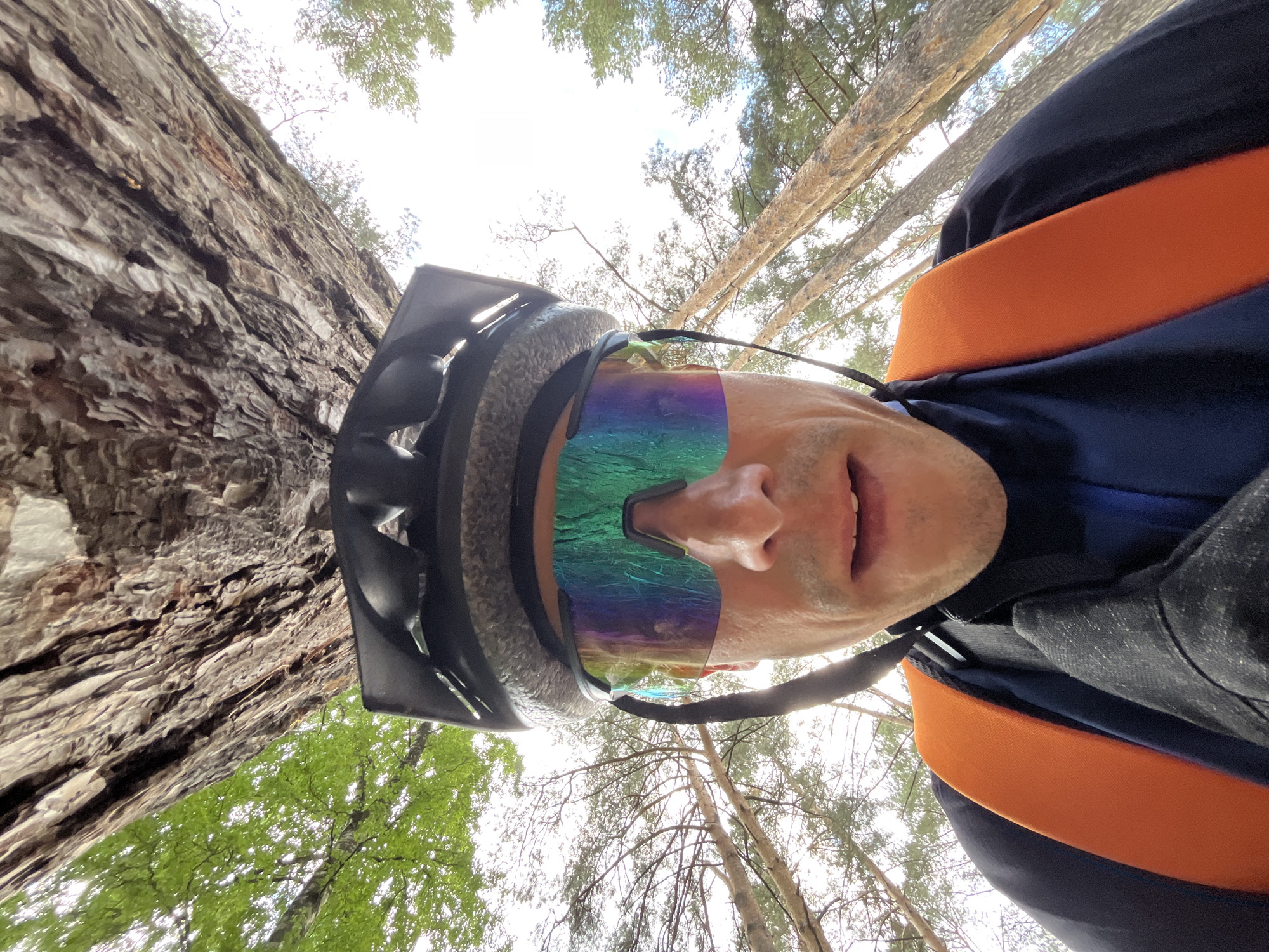 A person wearing a black helmet and iridescent cycling glasses is looking up at the camera from a low angle. They are wearing a blue cycling jersey with an orange backpack strap visible. The background consists of tall trees and the sky, suggesting an outdoor environment, likely a forest. The image is taken from a perspective that emphasizes the height of the trees. There is no specific indication of the city of Chaikovsky, Russia, within the visual elements of the image itself.
