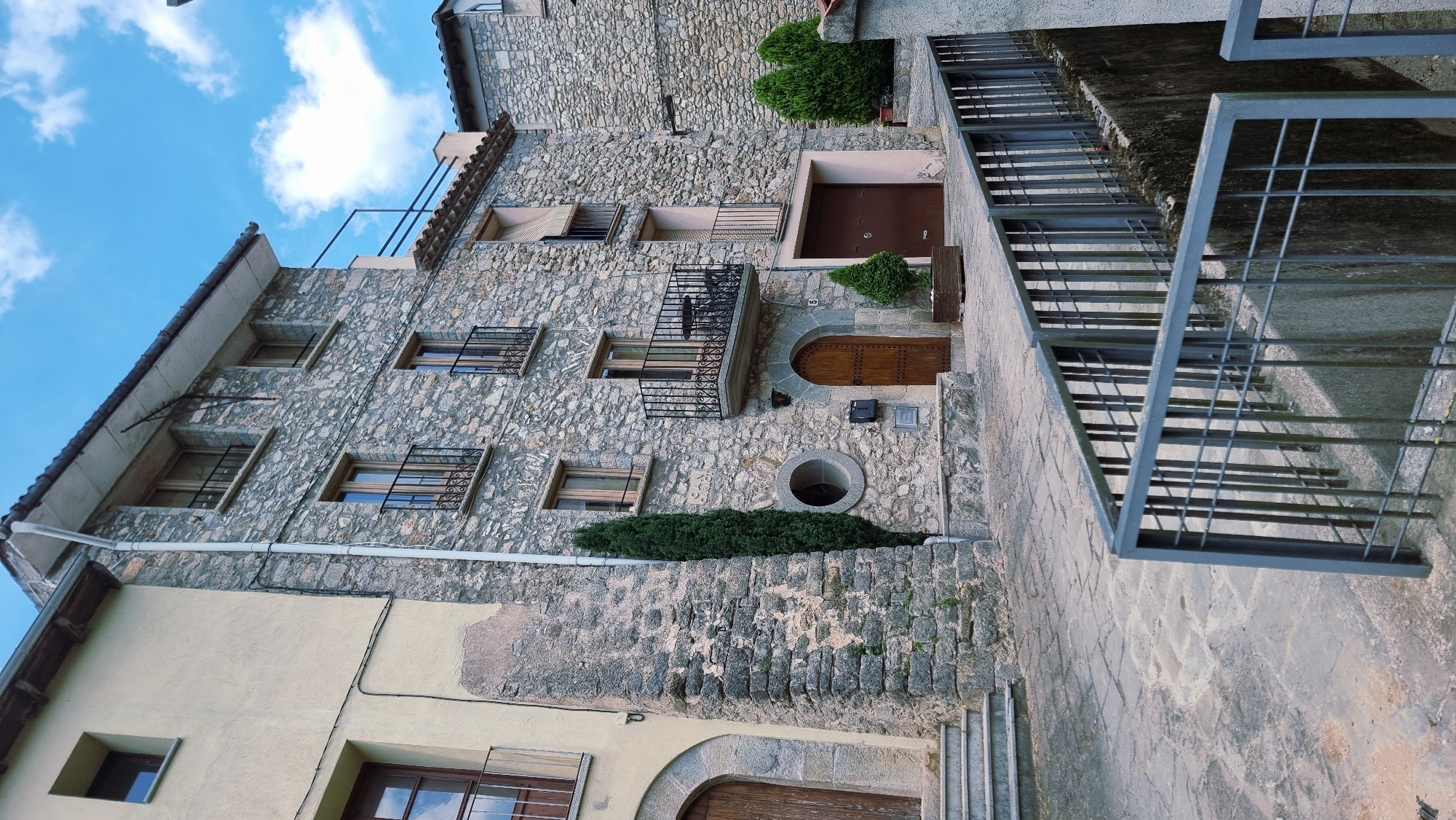 This image captures a side view of an old stone building with multiple windows, some featuring decorative metal grilles. The building appears to be made of rough-hewn stones, with a lighter colored section on the lower left. A dark brown door with a rounded arch is visible, as is a small, circular window. Green bushes are planted near the entrance and a balcony with a metal railing juts out from the upper floor.
To the right of the building, a gray metal staircase with railings ascends alongside a concrete structure. The stairs and the walkway are paved with stone. The sky above is blue with scattered white clouds, suggesting daytime. The scene is likely a residential area in a historic town, possibly in Montagut i Oix, Spain, given the architectural style. There are no people visible in the image. The overall impression is one of aged architecture and a quiet, perhaps rural, setting.