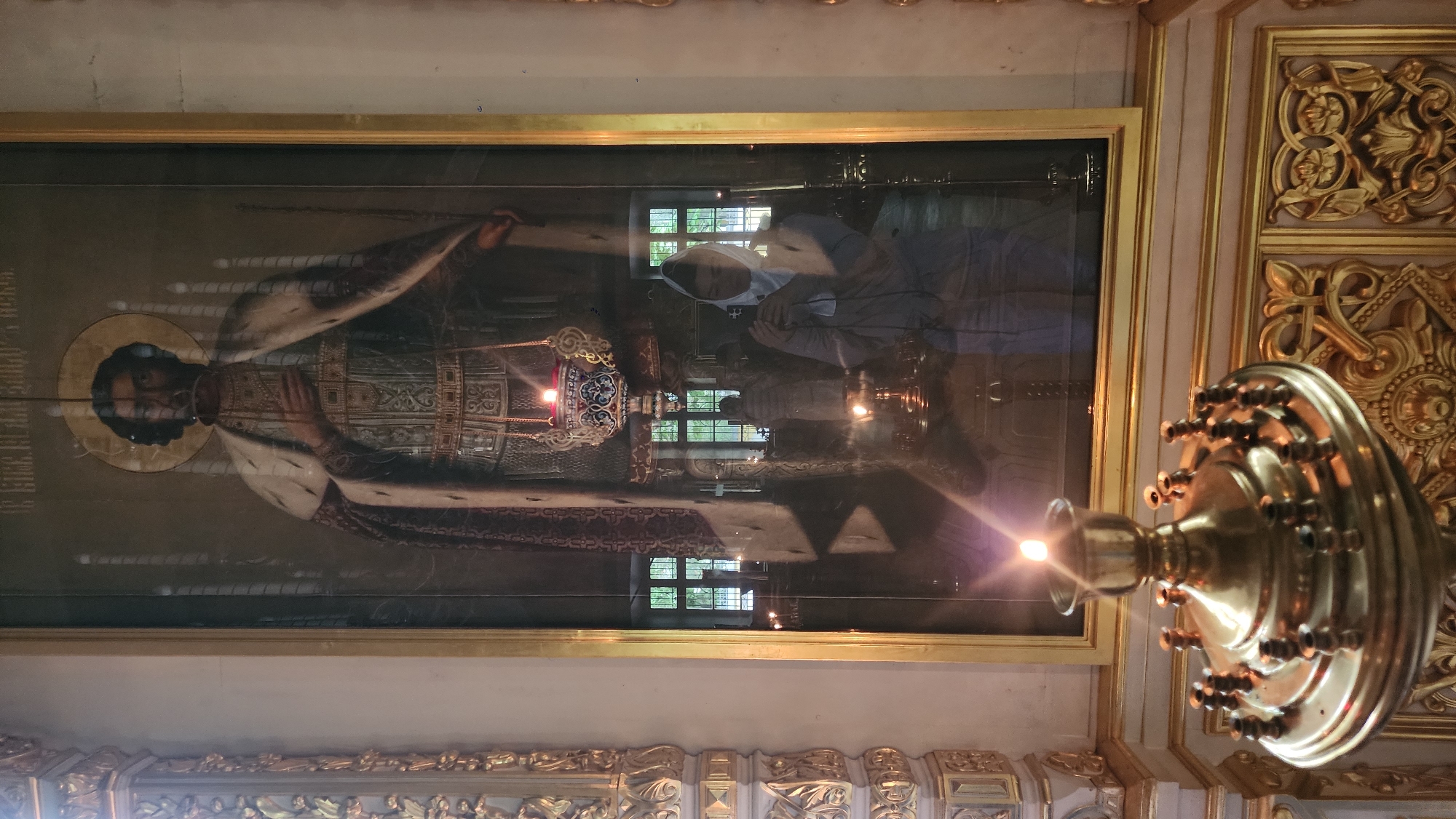 The image shows a framed painting hanging on a wall in what appears to be a building of religious significance. The painting depicts a man with a halo wearing regal attire, holding a scepter-like object. The reflection in the painting's glass shows a window with multiple panes and possibly other people. A gilded chandelier with numerous candle holders is visible in the foreground, casting a bright light. The surrounding architectural details, including the walls and ceiling, are heavily ornamented with gold-colored carvings. The location is Unknown.