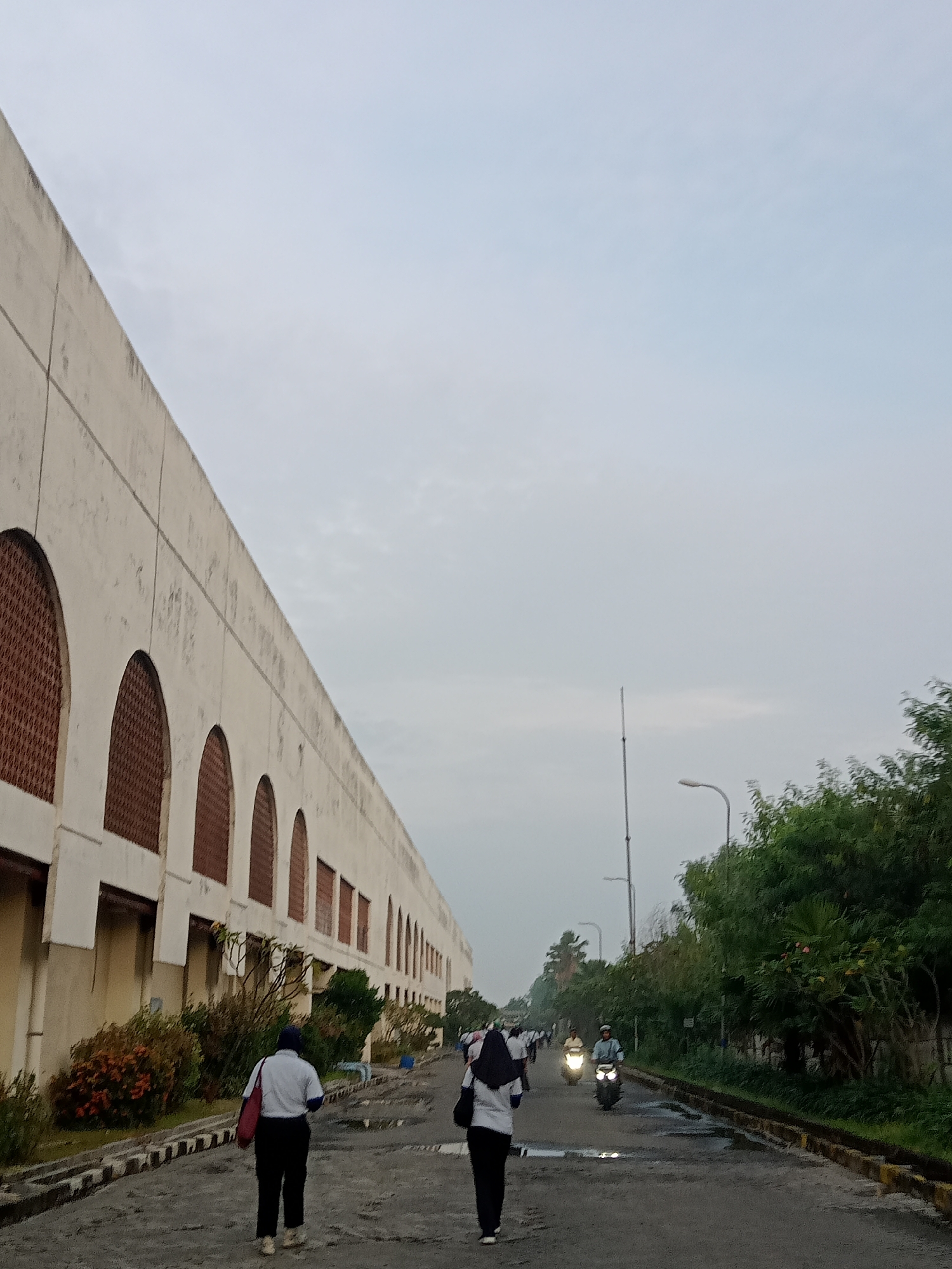 The image depicts a street scene in Sukomoro, Indonesia. The street is flanked by a long, low-rise building on the left, characterized by repetitive arched openings filled with what appears to be a patterned screen. On the right, a row of trees lines the street.
Two individuals are walking away from the viewer, dressed in what appears to be uniform-like clothing. Further down the road, there are more people and motorbikes with headlights on. Streetlights are visible intermittently along the roadside. The sky is overcast and the road surface appears damp. The general environment suggests an early morning or late afternoon setting.