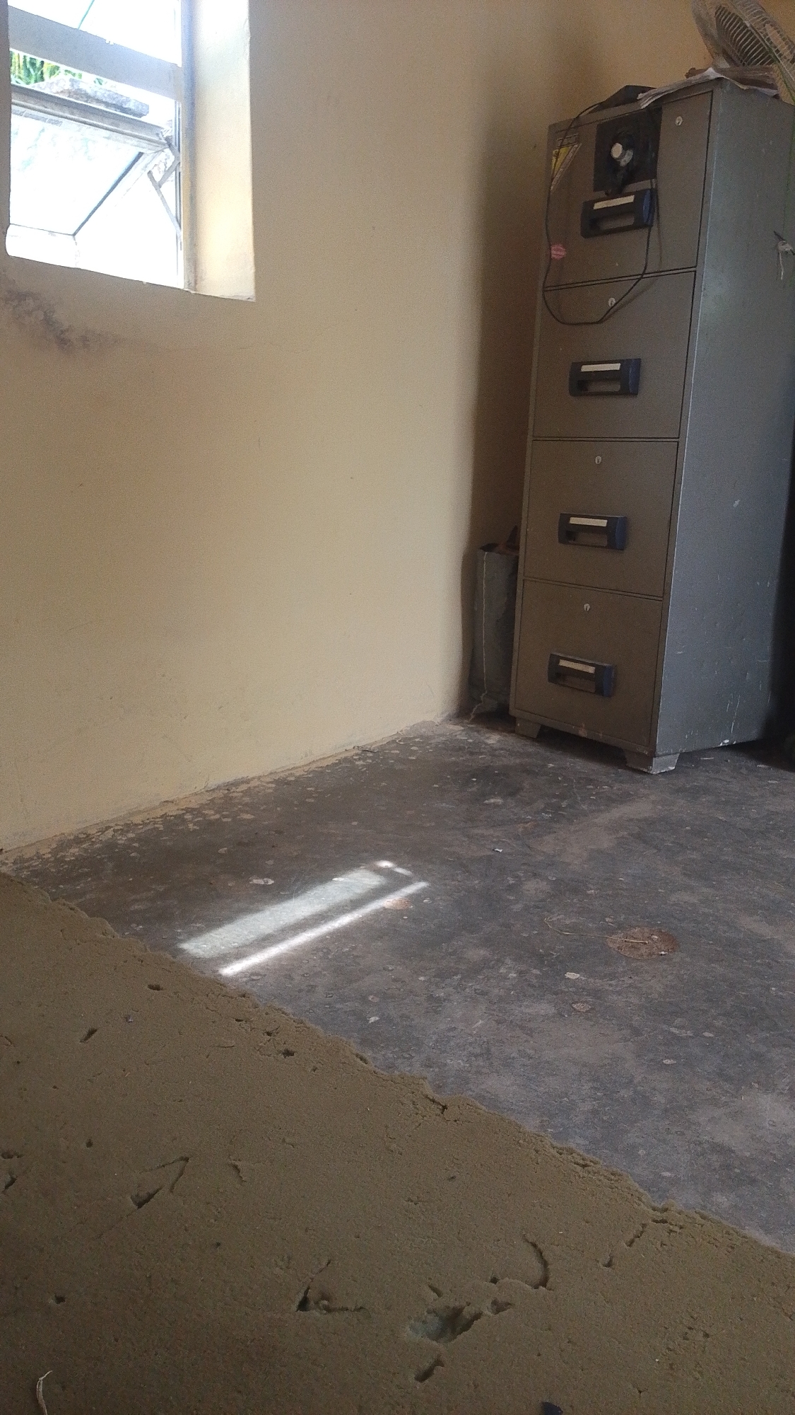 The image depicts an indoor scene, likely an office or storage room, located in Balewa, Nigeria. The room has pale yellow walls and a dark, concrete floor. Natural light streams in from a window on the left side of the frame, casting a distinct rectangular shadow on the floor. In the background, a tall, grey metal filing cabinet with four drawers stands against the wall on the right. A fan is visible at the top of the filing cabinet, and a thin black cord hangs from it. A rolled-up grey object, possibly a mat or fabric, is placed on the floor next to the filing cabinet. The foreground is occupied by a rough, sandy-colored surface, possibly a construction material or a poorly finished floor area, with several indentations. The overall impression is one of a functional, unadorned space.