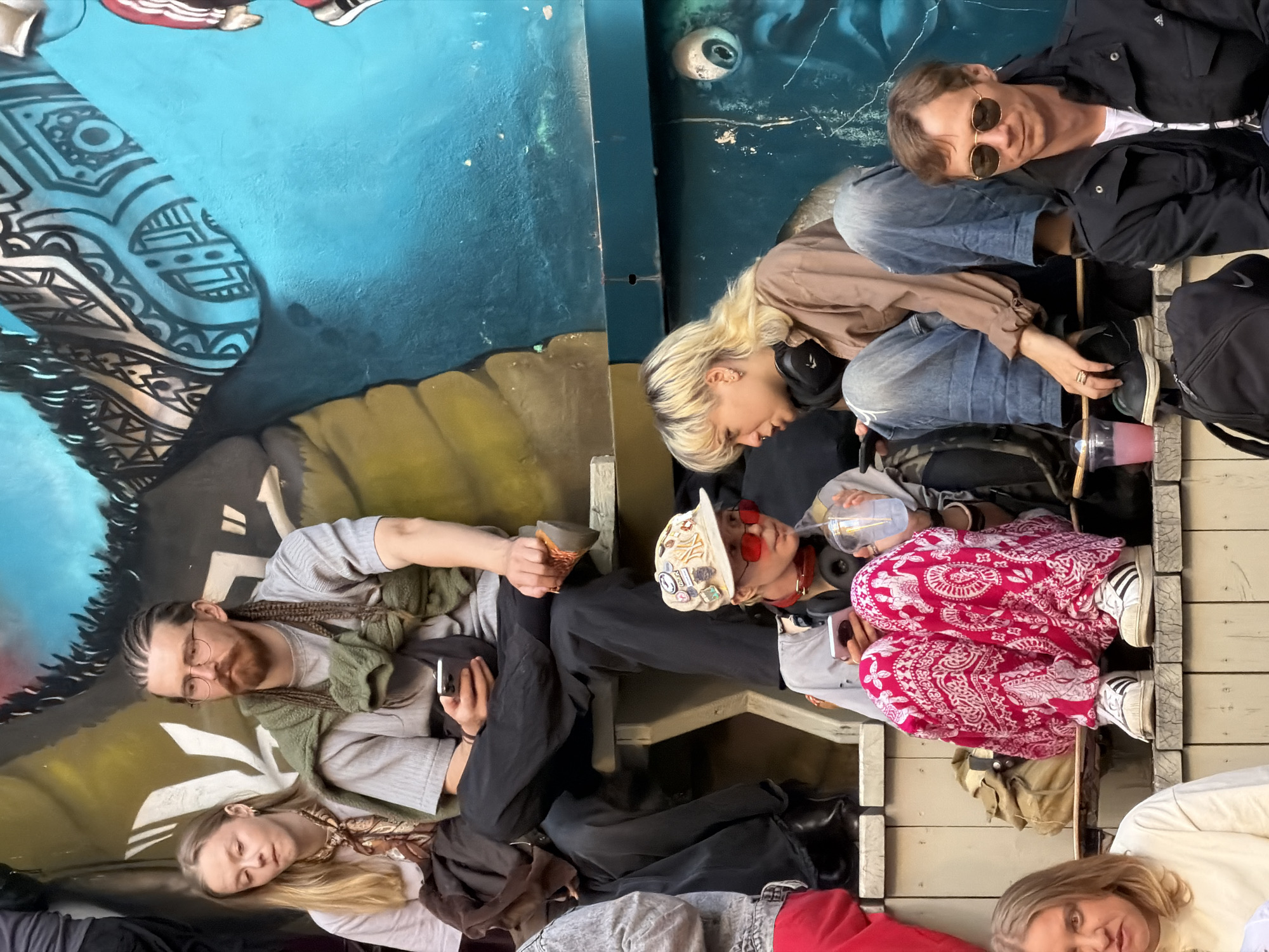 The image depicts a group of people sitting on wooden bleachers in front of a colorful graffiti-covered wall, suggesting an urban outdoor setting. On the left, a young woman with blonde hair wears a light-colored t-shirt and a patterned scarf. Next to her, a man with braided hair, a beard, and glasses is seated, holding a mobile phone. He wears a grey t-shirt with a green scarf draped over his shoulders. In the lower middle, a person with blonde hair and red-tinted sunglasses wears a baseball cap and a light-colored top with a red collar. They are holding a mobile phone and a clear plastic cup containing a beverage. To their right, another person with blonde hair, wearing headphones, is seated in a brown shirt and blue jeans, also holding a mobile phone. In the foreground on the right, a man with short brown hair and sunglasses wears a black jacket over a white t-shirt. He is looking down and appears relaxed. A black backpack is visible on the ground in front of him. The graffiti on the wall features blue, yellow, black, and white patterns, with a large eye-like motif visible on the blue section. The scene appears to be a casual gathering, possibly at an event or a public space. The presence of graffiti and the style of dress suggest a contemporary, urban environment. While no specific landmarks are identifiable, the overall aesthetic aligns with urban street art and gathering spots common in many cities.