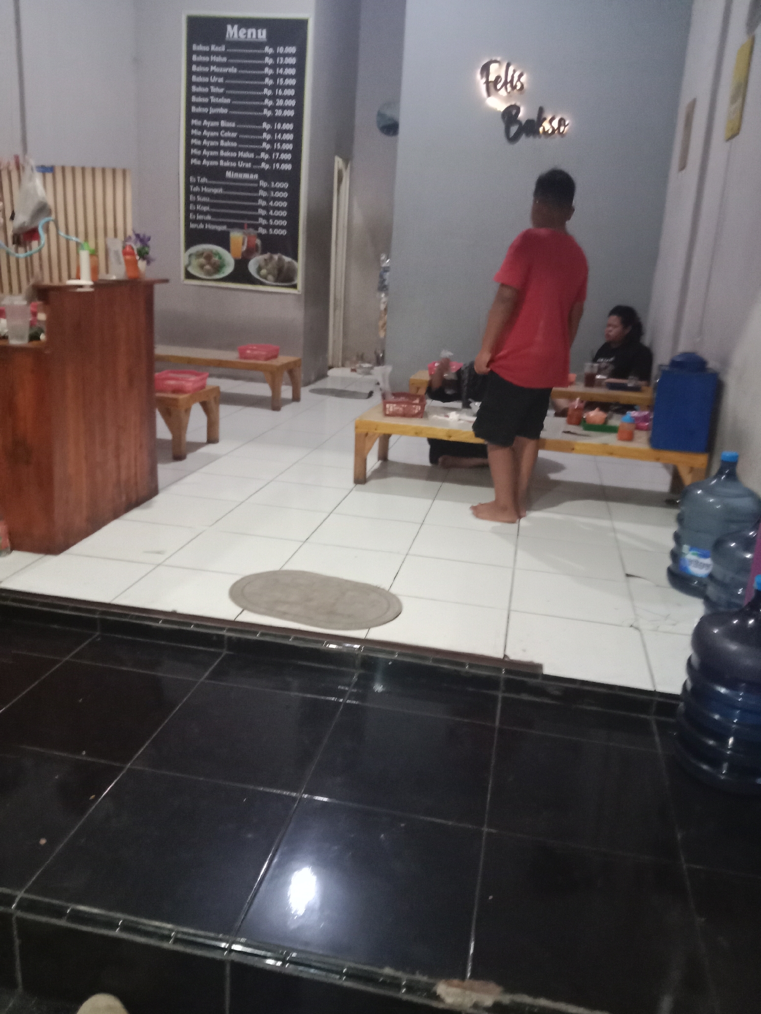 The image depicts the interior of a restaurant. A menu board is visible on the wall, listing food and drink items with prices in Indonesian Rupiah. The menu includes "Bakso Kecil" for Rp. 10.000, "Bakso Halus" for Rp. 13.000, "Bakso Mozarela" for Rp. 14.000, "Bakso Urat" for Rp. 15.000, "Bakso Telur" for Rp. 16.000, "Bakso Urotan" for Rp. 20.000, "Bakso Jumbo" for Rp. 20.000, "Mie Ayam Biasa" for Rp. 10.000, "Mie Ayam Ceker" for Rp. 14.000, "Mie Ayam Bakso" for Rp. 15.000, "Mie Ayam Bakso Halus" for Rp. 17.000, and "Mie Ayam Bakso Urat" for Rp. 19.000. Beverages listed are "Es Teh" for Rp. 3.000, "Teh Hangat" for Rp. 5.000, "Es Susu" for Rp. 4.000, "Es Kopi" for Rp. 4.000, "Es Jeruk" for Rp. 5.000, and "Jeruk Hangat" for Rp. 5.000. The restaurant has a simple setup with wooden tables and benches. A young person in a red t-shirt and black shorts stands facing away from the camera, while another person is seated at a table in the background. Large water dispensers are visible in the foreground and to the right of the image. The floor is tiled in white and black. A sign on the wall reads "Felis Bakso". The restaurant is likely located in Cangkring, Indonesia, as indicated by the presence of the menu in Indonesian and the style of the establishment.