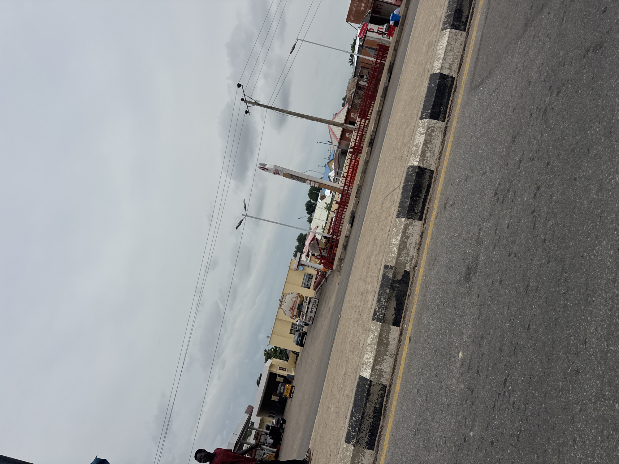 The image captures a view of a street scene in Jalingo, Nigeria, under an overcast sky. In the foreground, a grey asphalt road with a yellow line and a curb marked with alternating black and white blocks is visible. Beyond the curb, a paved area leads to several buildings.
To the left, a man in a red shirt and dark pants stands near a structure with a white roof, possibly a gas station. A yellow tricycle and a dark car are parked on the paved area. Further back, a yellow building with a sign that appears to say "Nigerwives" is prominent, with a red and white striped structure to its right. A tall, white pole with a sign on it stands near this building.
In the middle ground, more buildings and structures are visible, including some with red fencing. To the right, a building with a red roof and a sign that may read "Taman Usul" is present. People are seen in the distance near these establishments. Electrical poles and wires are strung across the sky. The overall environment suggests a commercial area within the city.