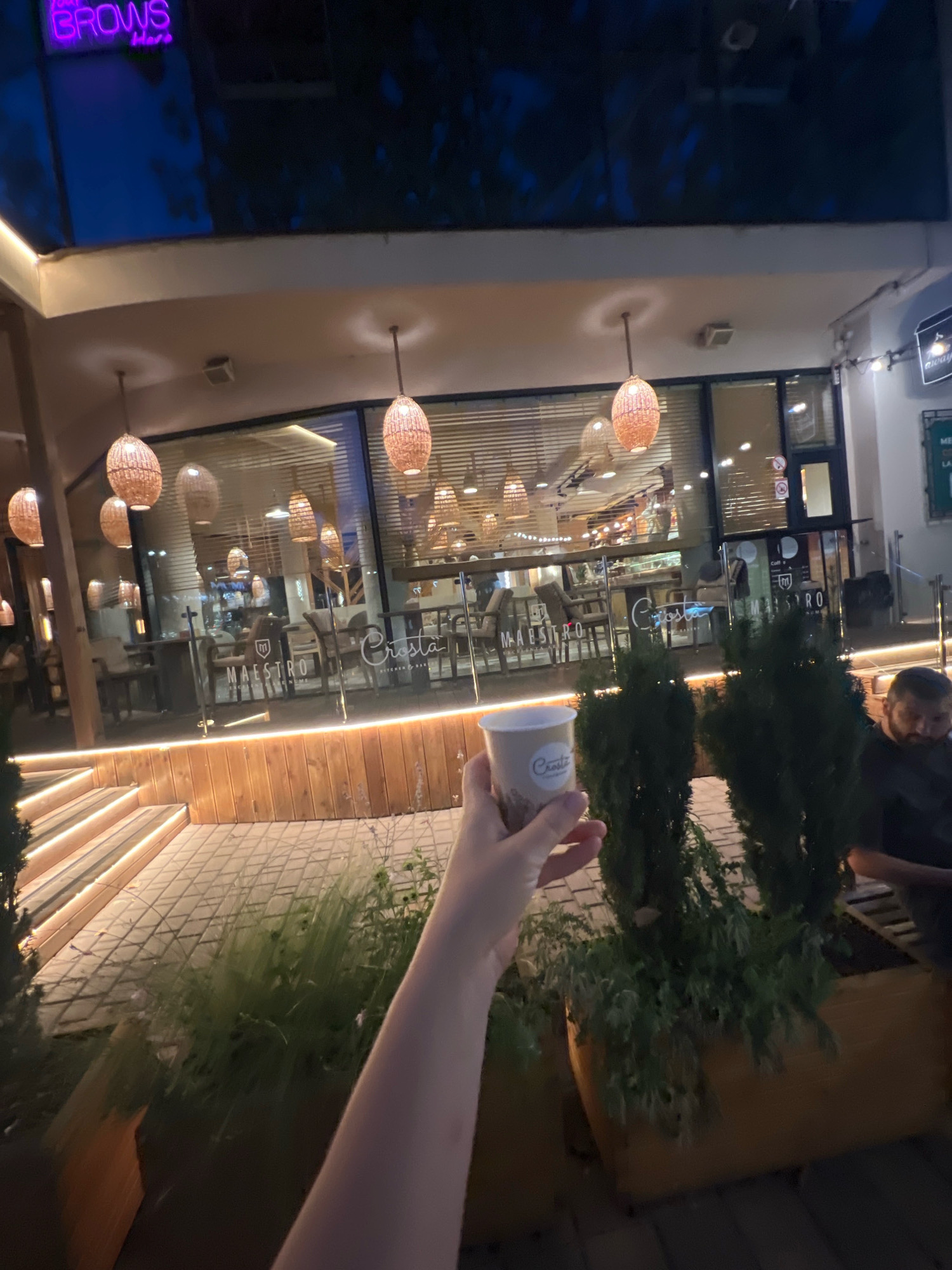 A first-person perspective shot shows a hand holding a paper coffee cup outside a restaurant. The cup has a logo on it, which reads "Croista". The restaurant's interior is visible through a glass window, showing tables and chairs. Several woven pendant lights hang from the ceiling of the restaurant, which also has a neon sign that says "BROWS". A man is visible in the background, sitting at a table. The ground is paved with square tiles, and there are planters with greenery. Wooden stairs with embedded lights are to the left of the frame. The overall scene appears to be an outdoor seating area of a restaurant at dusk or night.