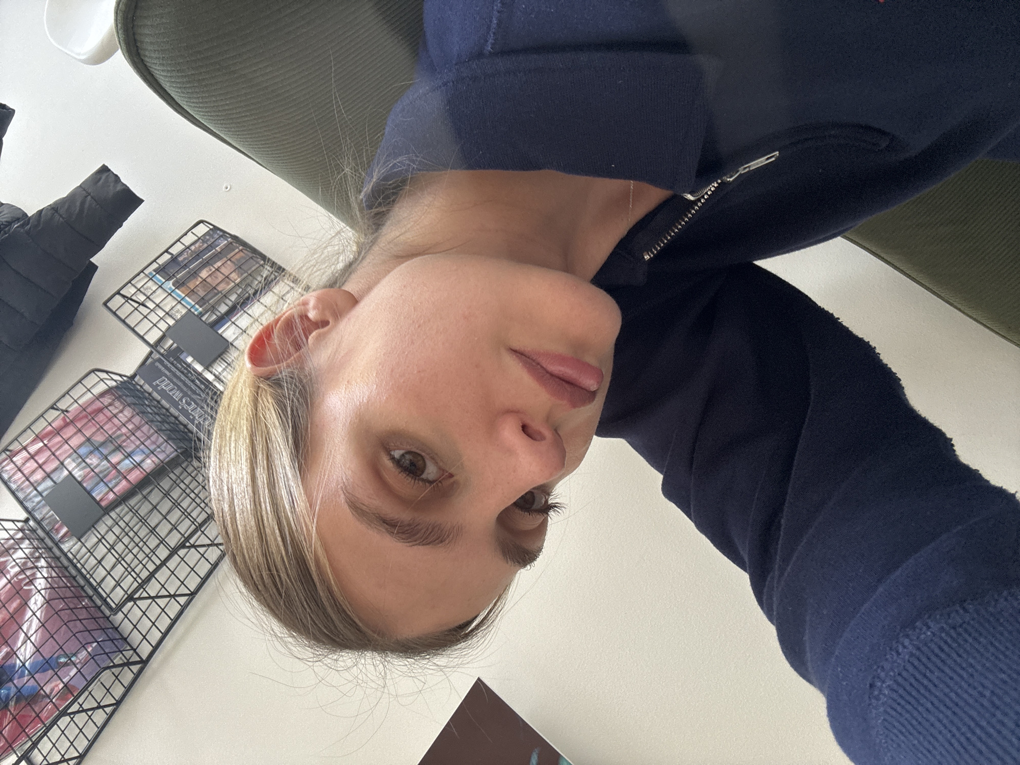 A person with fair skin, blonde hair, and brown eyes is looking at the camera. They are wearing a dark blue zip-up sweater. In the background, there is a metal magazine rack, a dark-colored coat, and a green cushioned chair. The setting appears to be indoors, with plain white walls. The city and precise location are unidentifiable.