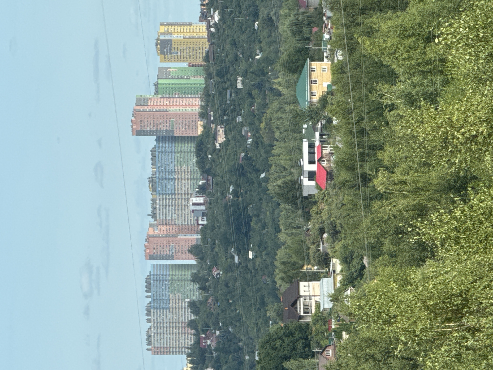 The image captures a panoramic view of the city of Ankudinovka, Russia, showcasing a striking contrast between modern high-rise buildings and traditional low-rise structures nestled amidst lush greenery. In the background, a row of colorful apartment complexes stands tall, their facades adorned in shades of blue, pink, green, and yellow, indicating a modern residential development. The mid-ground features a sprawling landscape of rolling hills covered in dense, vibrant green trees, punctuated by a scattering of smaller houses and buildings, suggesting a suburban or semi-rural environment.
The foreground is dominated by a thick canopy of trees, their leaves a lively green, hinting at a summer or late spring season. Interspersed within the foliage are glimpses of more houses, some with red roofs, and what appear to be power lines stretching across the scene, signifying the presence of infrastructure. A few visible structures include a white building with a dark roof, a yellow house with a green roof, and a smaller house with a red roof. The sky above is a clear, pale blue with a few wispy clouds, bathing the entire landscape in natural light. No people are visible in the image. The overall impression is one of a developing urban area integrating modern architecture with natural landscapes.