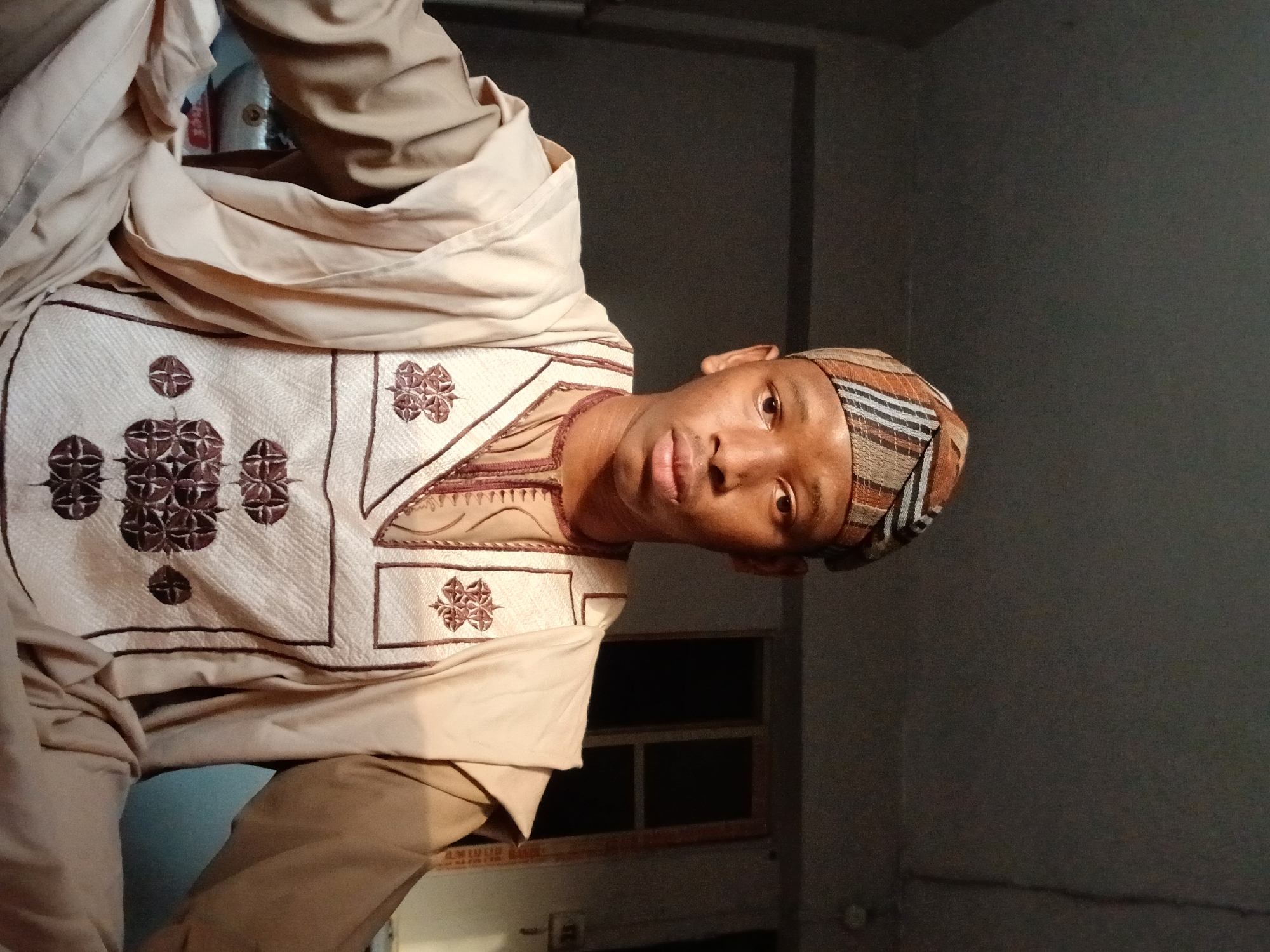 A man is indoors, wearing a light brown and white traditional outfit with dark brown embroidery. He is also wearing a striped head covering. The background consists of a dark wall and a window or door. The location is unknown.
