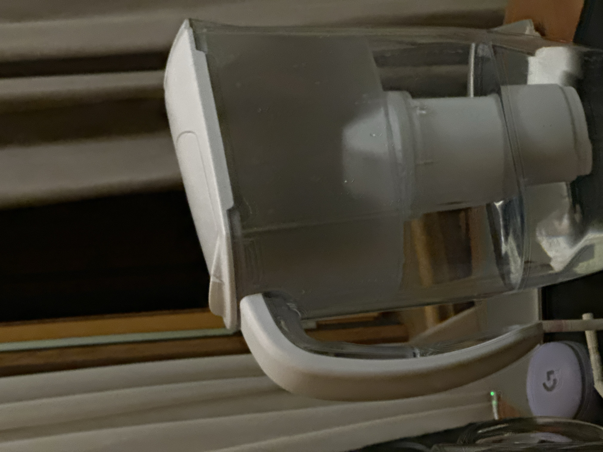 A close-up shot focuses on a clear water filter pitcher with a white handle, situated indoors in an unidentified location. The pitcher appears to be partially filled, and its filter component is visible inside. It rests on a surface near a set of horizontal blinds that are out of focus in the background. In the lower right corner, a portion of a Logitech device is visible. The lighting is soft, and the focus is sharpest on the pitcher's front.