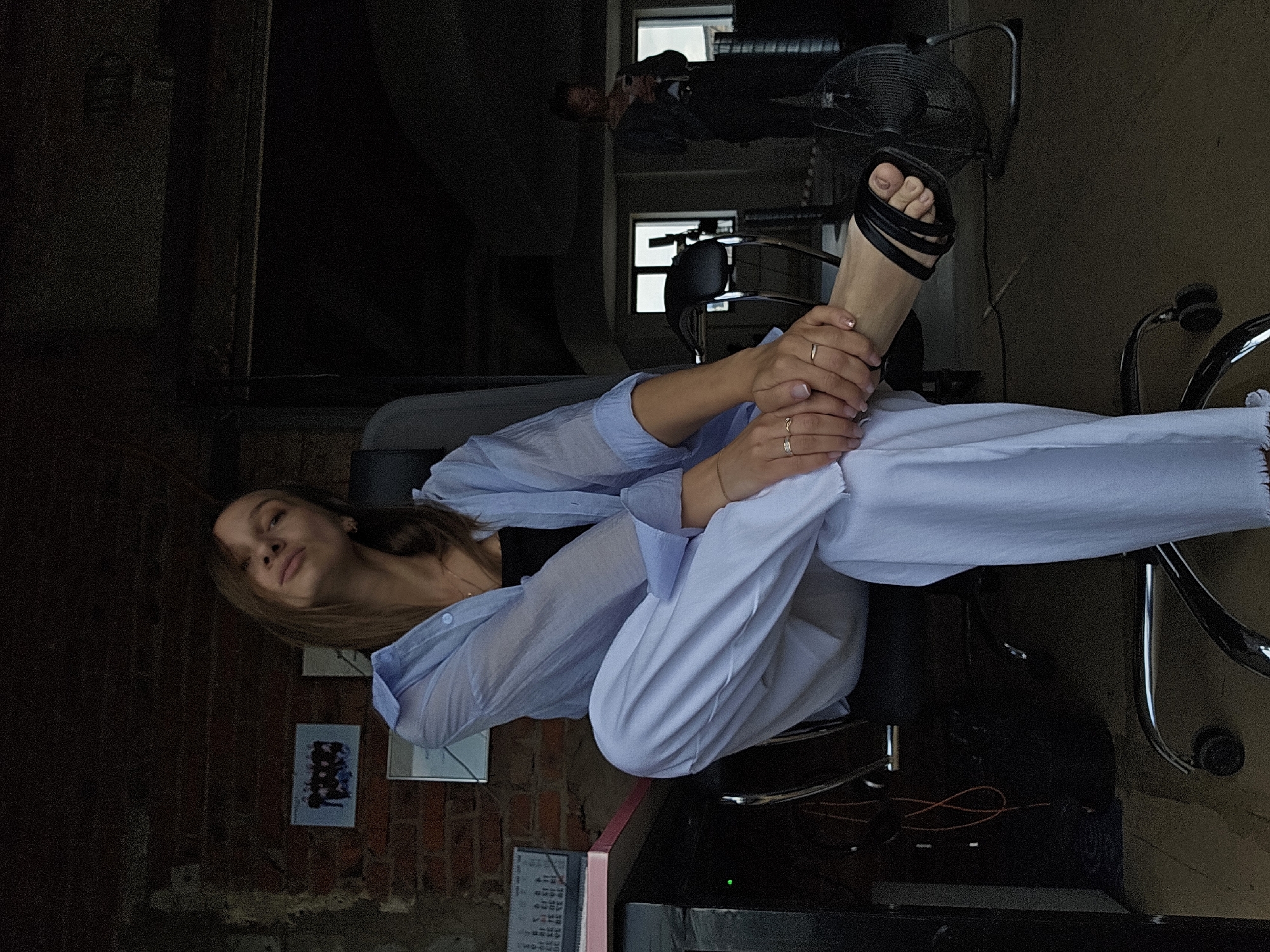 A woman is seated in a black office chair in a room with exposed brick walls. She is wearing a light blue button-down shirt over a black top and white pants, with black sandals on her feet. Her legs are crossed, and she is looking upwards with her head tilted back. There are several framed pictures on the wall behind her. A desk is visible to her left, with a pink notebook and a calendar on it. A black fan is positioned to her right. In the background, another woman is standing and looking at her phone. The lighting suggests it is either early morning or late evening. The overall environment appears to be an indoor workspace or studio.