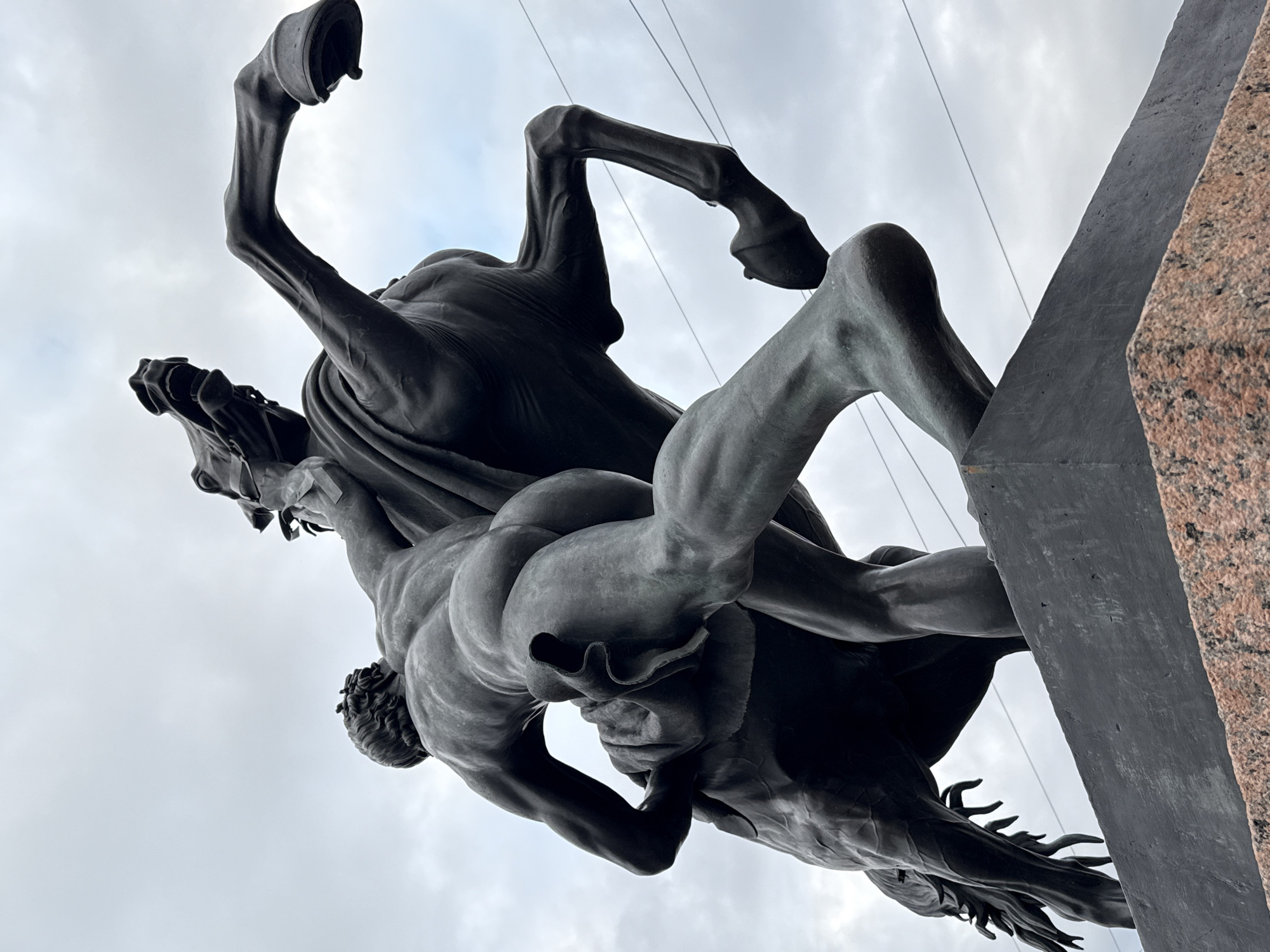 The image captures a low-angle, dramatic view of a bronze equestrian sculpture in Saint Petersburg, Russia, likely depicting the Bronze Horseman. The sculpture portrays a muscular nude male figure wrestling with a rearing horse. The man is positioned on the horse's back, his body contorted as he grapples with the animal's reins and head. The horse is depicted in a powerful, dynamic pose, its front legs raised and its mouth open in a silent struggle. The sculpture is mounted on a dark, angular pedestal, which itself rests on a foundation of reddish-pink granite. The background is dominated by an overcast, pale gray sky with some subtle blue tones, suggesting a cloudy day. Thin wires, possibly power lines, stretch across the sky in the upper right quadrant of the image. The lighting is diffused due to the cloud cover, casting soft shadows that highlight the musculature of the figures and the texture of the bronze. The perspective emphasizes the scale and grandeur of the monument.