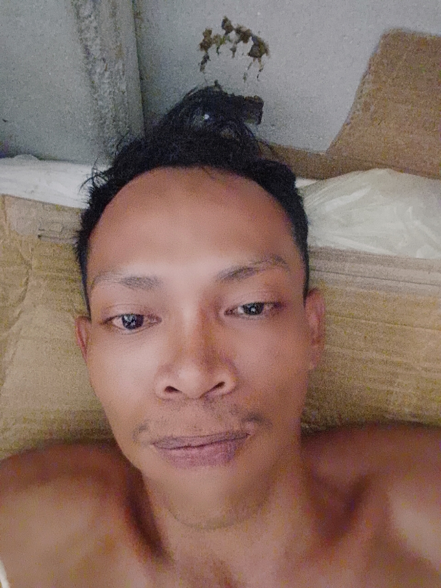 A man is lying on a pillow. The location is Bukit Ketok, Indonesia, as specified. The man has dark hair and dark eyes. There is staining visible on the ceiling in the background. The pillow has a white pillowcase on top of a tan patterned pillow. The man appears to be topless.