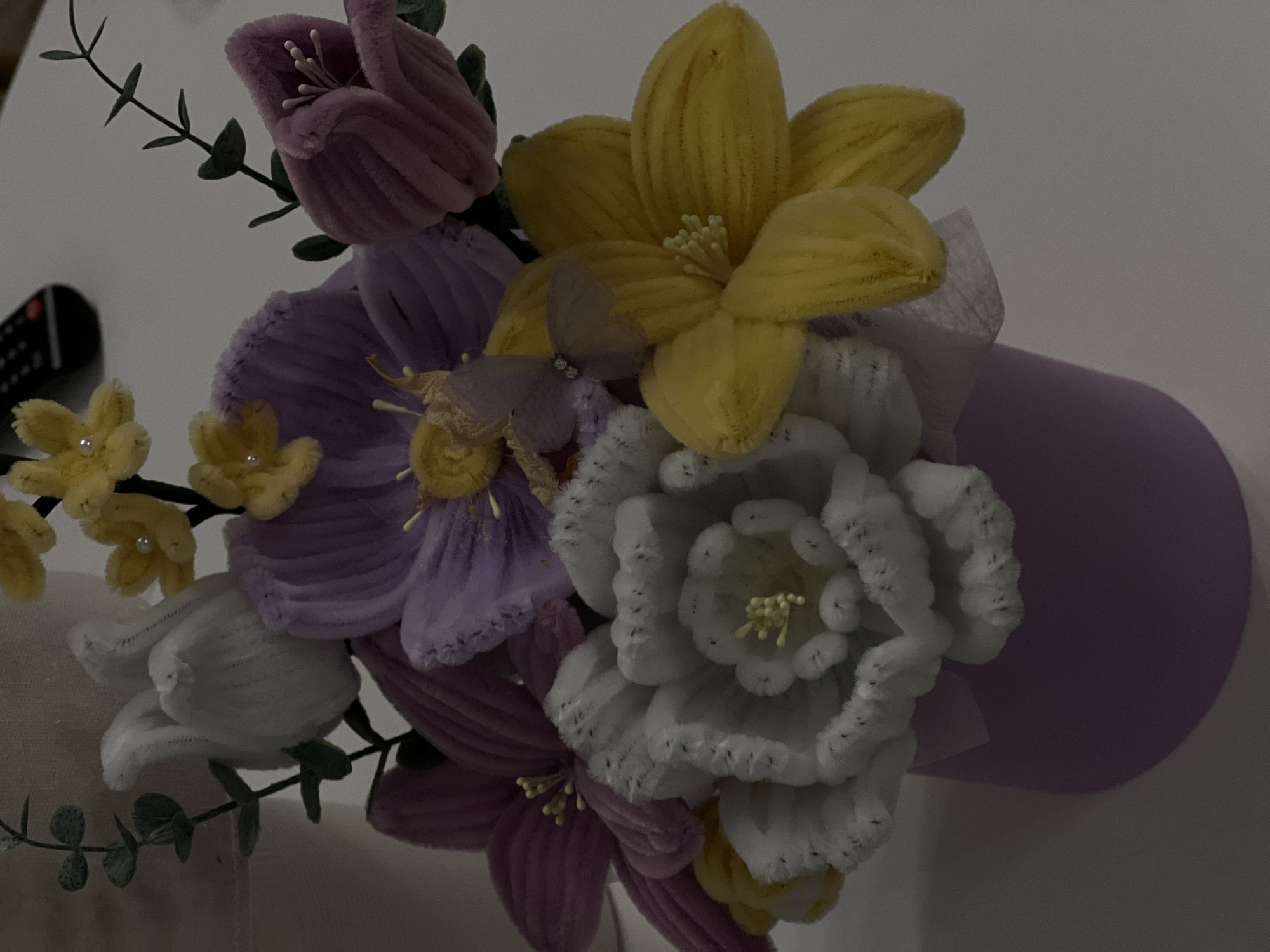 The image displays a close-up of a bouquet of artificial flowers in a lavender pot. The flowers are crafted from a soft, fuzzy material, resembling pipe cleaners, and are arranged in various colors including white, purple, pink, and yellow. Some of the flowers have delicate stamen-like centers. Green artificial leaves are interspersed among the blooms, adding a touch of greenery. A small, translucent butterfly decoration with beaded wings is attached to one of the purple flowers. The arrangement is set against a neutral, dimly lit background, with a hint of a lamp or light fixture visible on the left. The composition suggests an indoor setting, possibly a home or a craft display. There is no indication or context related to Florești, Romania, within the visual information of the image.