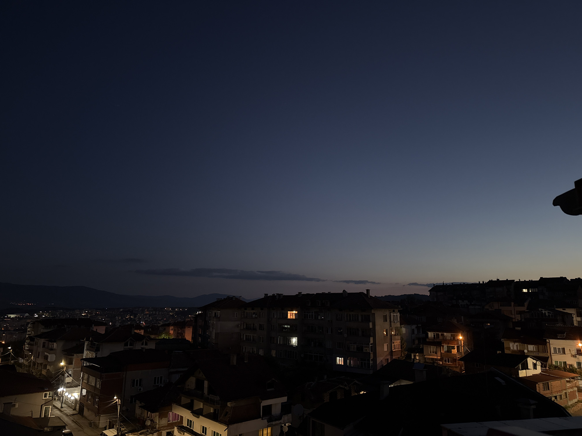 The image depicts a dusk or dawn scene over a residential area in Blagoevgrad, Bulgaria. The sky is a deep gradient of dark blue and lighter hues near the horizon. Silhouetted mountains are visible in the distance. The foreground and midground are filled with buildings, predominantly multi-story apartment blocks and houses, with varying rooflines and architectural styles. Many windows are illuminated, indicating that people are inside their homes. Streetlights are on, casting pools of light on the streets and illuminating some of the building facades. A few cars are visible parked on the street. The overall impression is a quiet urban evening or morning in a city.