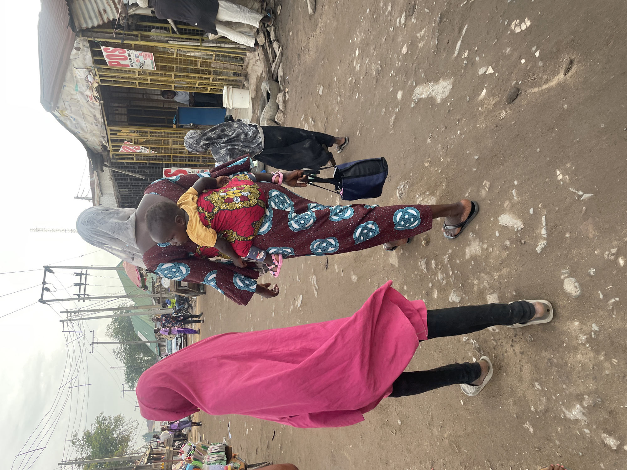 The image captures a vibrant street scene, likely a bustling market or commercial area in Abuja, Nigeria, under bright, possibly overcast, daylight. The setting is characterized by an unpaved, dusty street with scattered small rocks and debris.
In the foreground, a woman wearing a colorful patterned dress (red, blue, and white) carries a baby securely on her back. The baby is dressed in a yellow top. This woman is also seen wearing sandals and holding a blue shoulder bag. Walking beside her is another individual dressed in a long, flowing pink garment over black pants, with white sandals. Further into the midground, another person in dark clothing and a head covering walks away from the viewer.
The background on the left features informal market stalls or shops constructed with corrugated iron roofs and yellow metal grate facades. A prominent red and white sign on one of these structures clearly reads "POS AVAILABLE," indicating the presence of point-of-sale services. A person can be seen inside one of the stalls. Overhead, numerous power lines and utility poles crisscross the sky, with trees and distant structures visible further down the street. The overall scene depicts everyday activity in a lively urban environment.