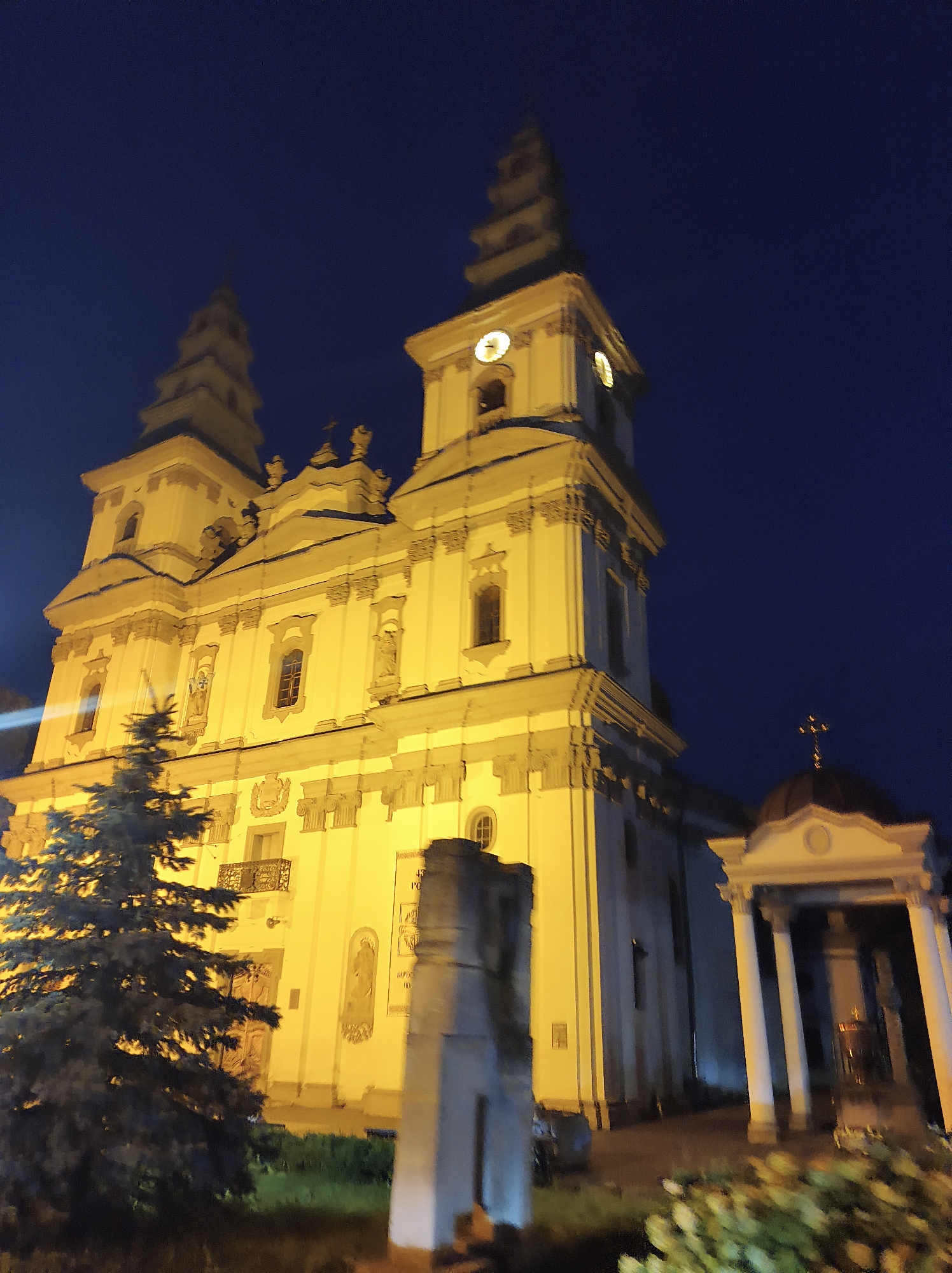 The image shows a large, yellow-lit church at night. The church has multiple towers, some with spires, and a clock face visible on one of the towers. In front of the church, there is a dark evergreen tree and a stone structure. To the right of the church, there is a small building with columns and a cross on top. The sky is dark blue, indicating nighttime. This is the Ternopil Theological Seminary, located in Ternopil, Ukraine.