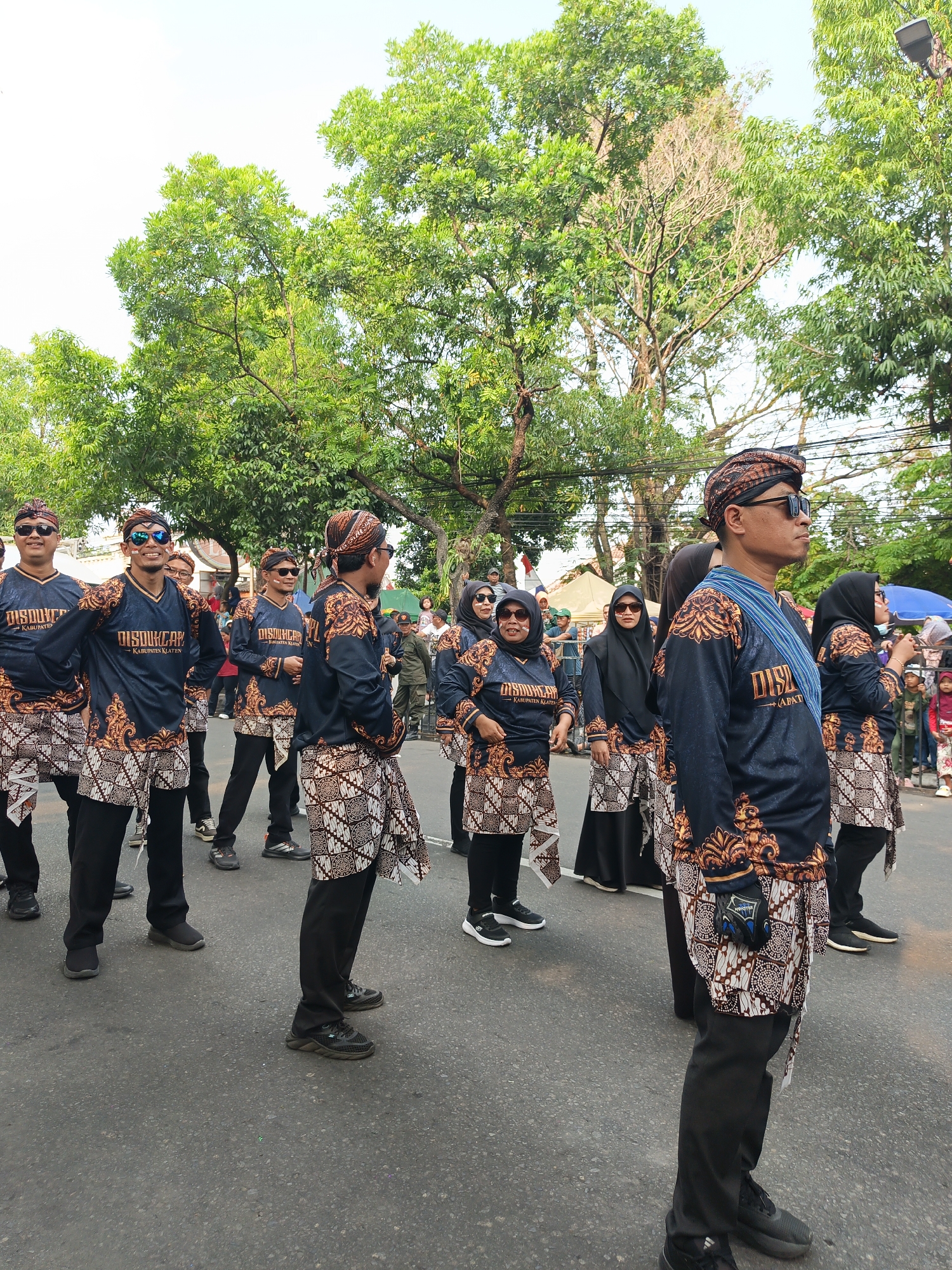 In Gayamprit, Indonesia, a group of people are participating in a parade. They are dressed in matching dark blue shirts with intricate gold and white batik patterns on the sleeves and collars, and matching batik sarongs worn over black pants. Many of the individuals are wearing sunglasses and traditional headwear. They appear to be walking in formation, some with their hands on their hips and others with their arms relaxed at their sides. The background shows lush green trees lining the street, with a clear sky above. There are some spectators visible in the distance, and tents can be seen set up along the side of the road. The scene suggests a festive community event.