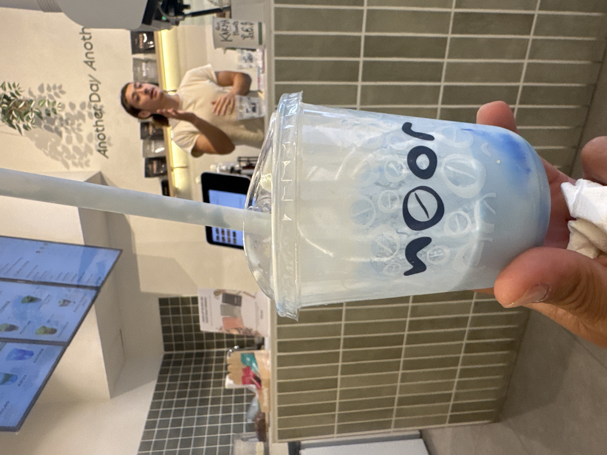 The image captures a casual scene inside a modern cafe in Barcelona, Spain, oriented sideways from its original perspective. In the foreground, a person's hand, holding a white tissue, clutches a clear plastic cup containing a layered blue and white beverage, possibly a bubble tea or a specialty drink. A white straw protrudes from the cup, which is adorned with the prominent black text "SOOR" and subtle circular patterns.
In the background, another individual, likely a barista or staff member, stands behind a counter. This person has dark hair and is wearing a light-colored top, appearing engaged in an activity at the counter. Behind them, a white wall features the partial text "Another Day Anoth" and the shadow of a plant. Further back, shelves display various products, and a small sign with text like "KARA" and "Beans" with a price is visible. The wall to the right of the counter is covered in horizontal, light brown/tan tiles, and a large digital menu screen displaying various drink options is partially visible on the left. The overall setting suggests a contemporary and welcoming atmosphere within the cafe.