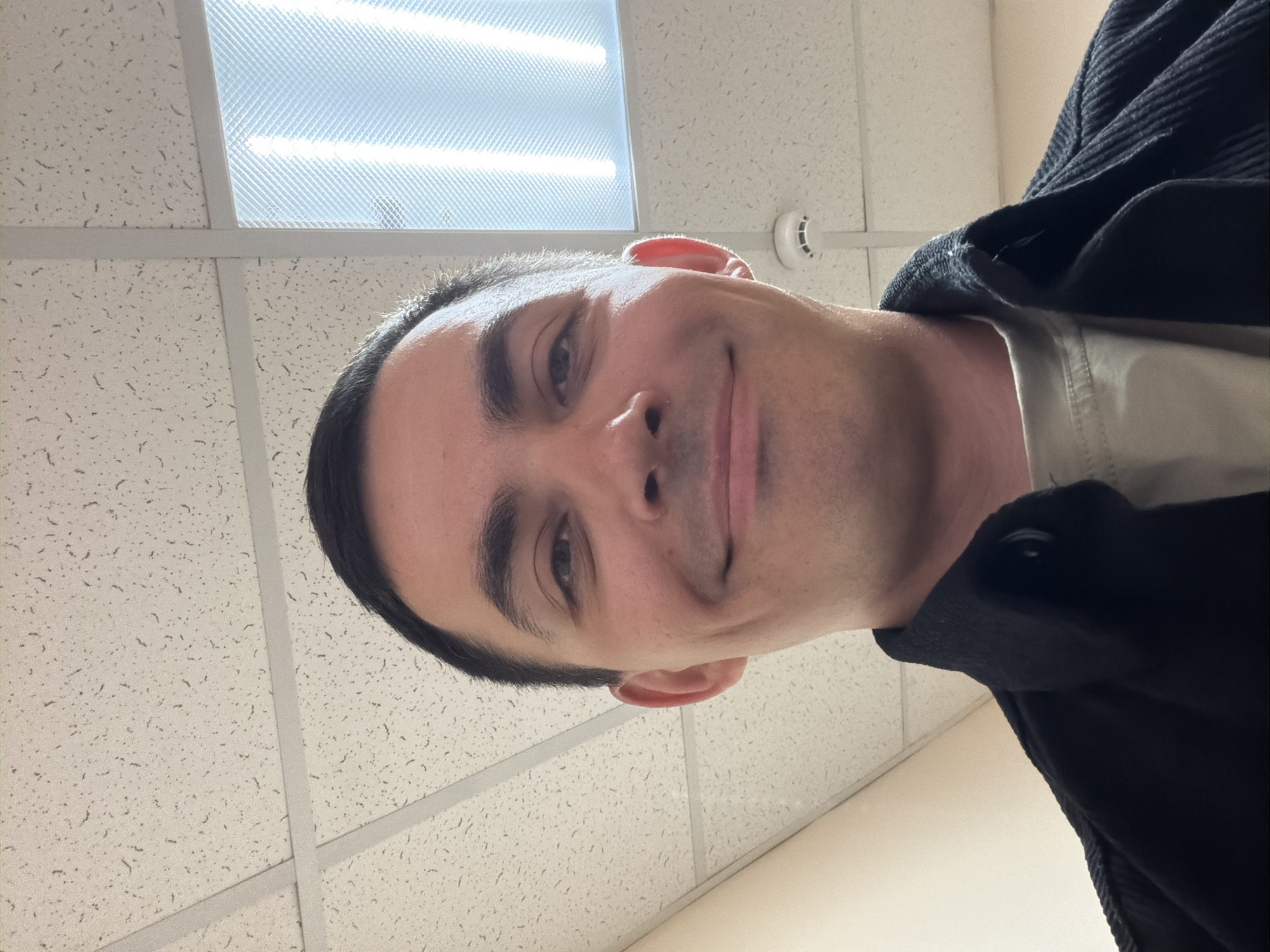 A man with short, dark hair is looking directly at the camera with a slight smile. He is wearing a dark jacket over a light-colored shirt. The background is a drop ceiling with fluorescent lighting panels. A round, white object, possibly a smoke detector, is visible on the ceiling. The scene is brightly lit. The location is not discernible from the image.