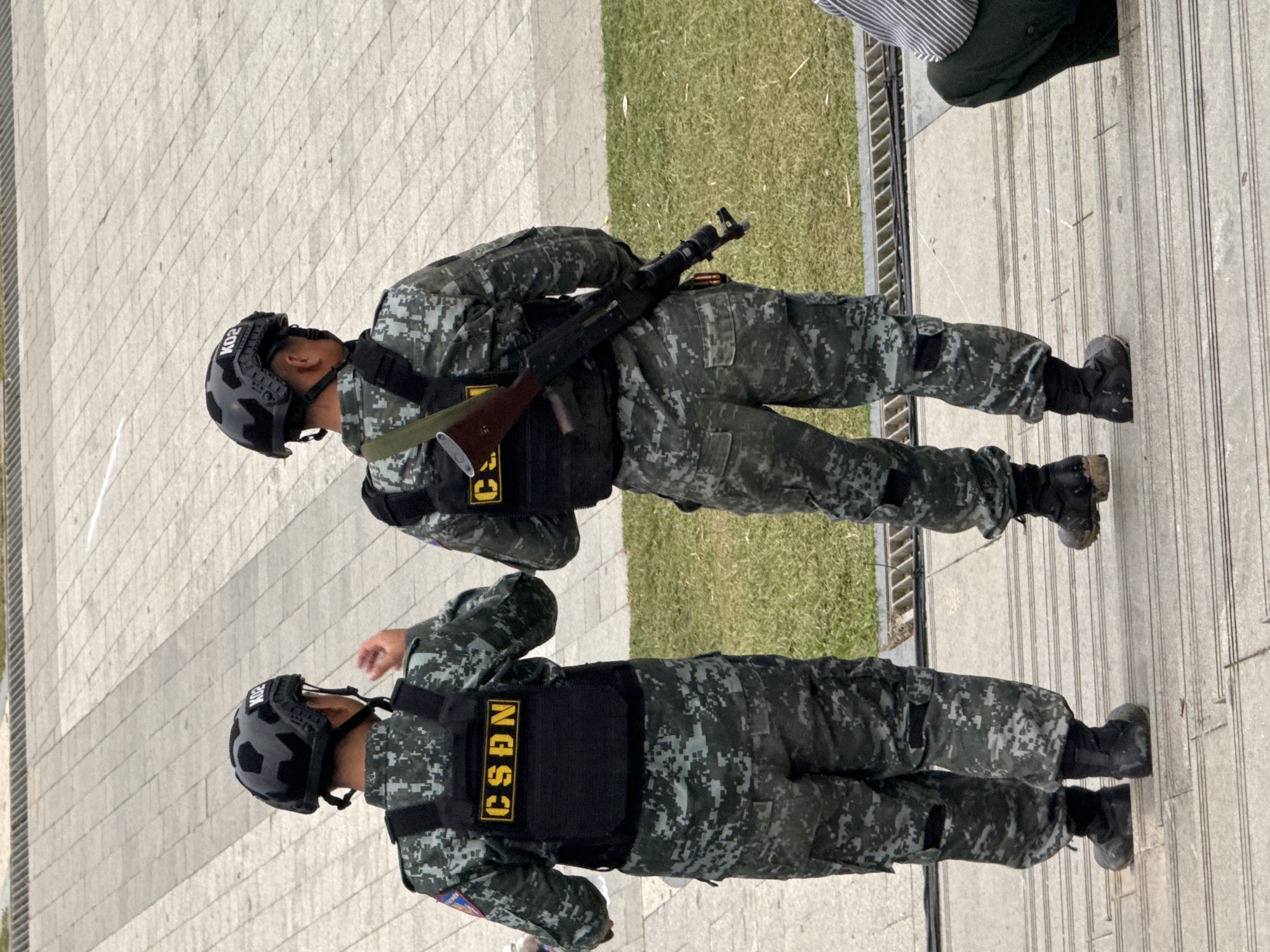 Two individuals in military-style uniforms stand on a paved walkway. Both are wearing camouflage patterned clothing, tactical vests, and helmets. The person in the foreground has "CSDN" clearly visible in yellow lettering on their vest. The person behind them also has lettering on their vest, but it is partially obscured. The individual in the background is holding a rifle. The setting appears to be an outdoor public space with a grassy area to the right and a textured walkway. The lighting suggests it is daytime. The overall scene conveys a sense of security or preparedness. The image is oriented vertically.