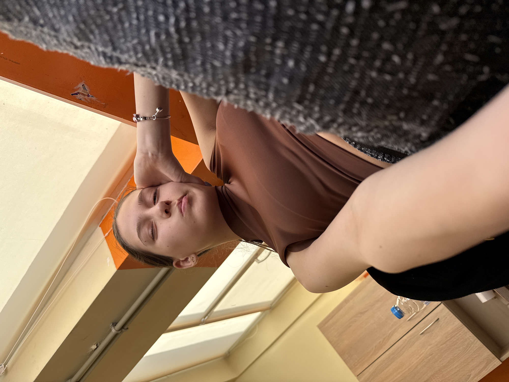 A young woman in a brown shirt is seen close-up in an indoor setting, possibly a room in Sofia, Bulgaria, given the request. She is positioned with her face and torso visible, but her orientation is such that the background appears skewed, suggesting the camera is at an odd angle. The woman has fair skin and is holding her left hand near her head. She seems to have her eyes closed.
The background includes elements of a room, such as a window, a light orange beam, some type of couch with a grey and black fabric, and a cabinet with a plastic bottle on top. The lighting appears to be natural, possibly from daylight entering the window.