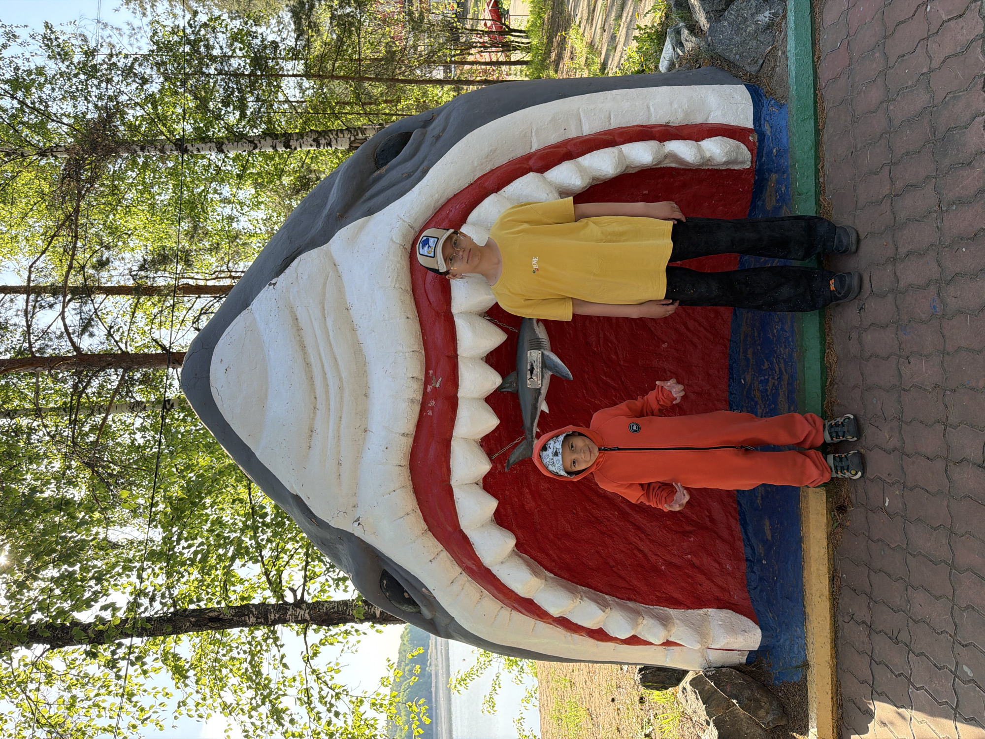 Two children are lying inside the open mouth of a large, concrete shark statue. One child, wearing a yellow shirt and a baseball cap, is positioned near the top of the mouth, while the other child, wearing an orange jumpsuit and a bandana, is at the bottom. Inside the shark's mouth, a smaller model shark is also visible. The shark statue is painted with gray, white, and red. The statue is placed on a red brick walkway next to some trees. The picture was taken in the city of Chaykovsky, Russia.