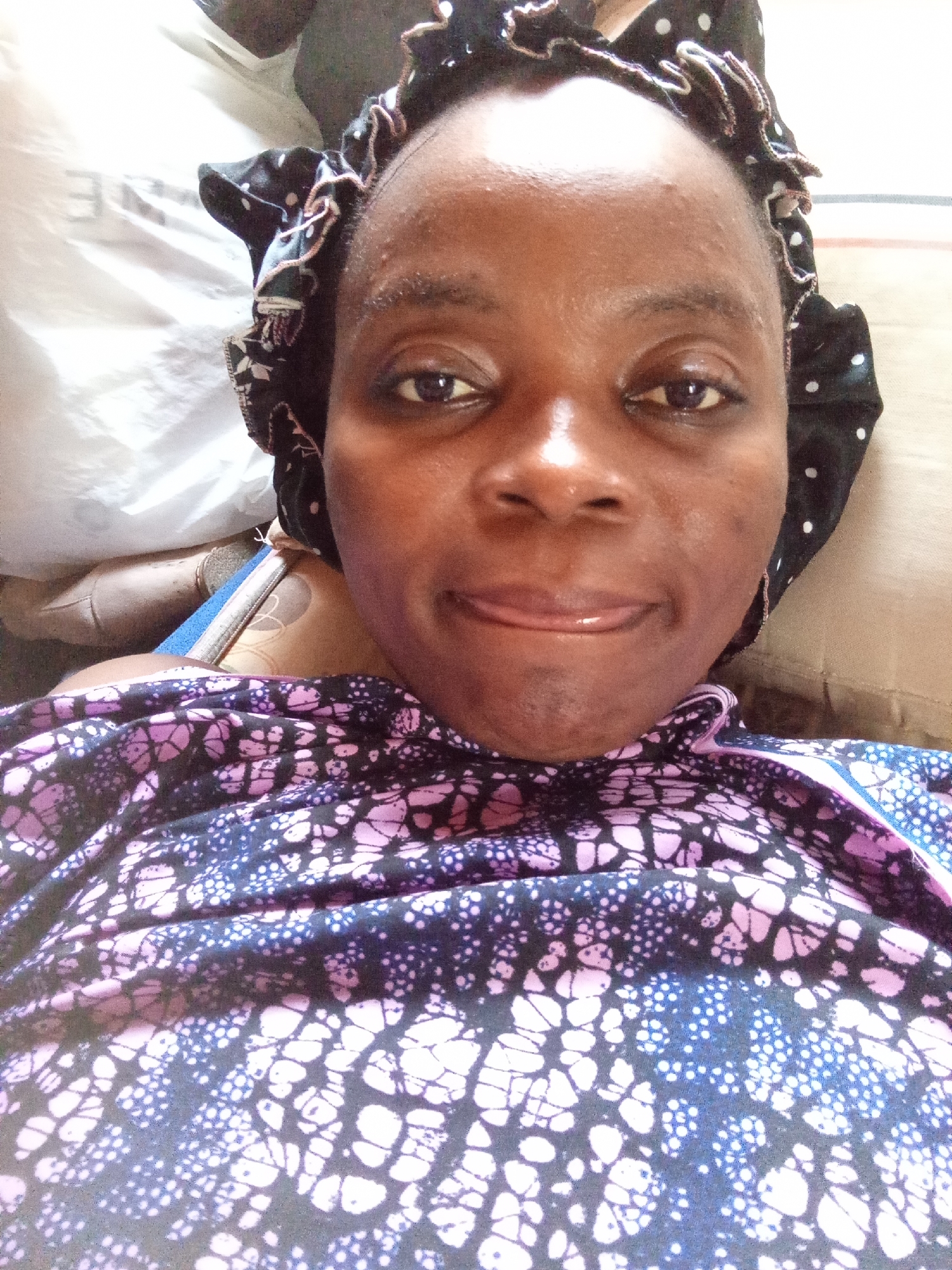 A close-up, overhead shot shows a dark-skinned woman wearing a black and white polka dot bonnet and a purple and blue patterned top. Her face is turned towards the camera, with her lips slightly parted. She appears to be lying down on a surface that is partially obscured by her body and clothing. The background consists of a light-colored fabric, possibly bedding, and a portion of what appears to be a person's leg covered in khaki pants. The setting is indoors, and the lighting suggests it is daytime. The specific location within Kason, Nigeria, is not discernible from the image itself.