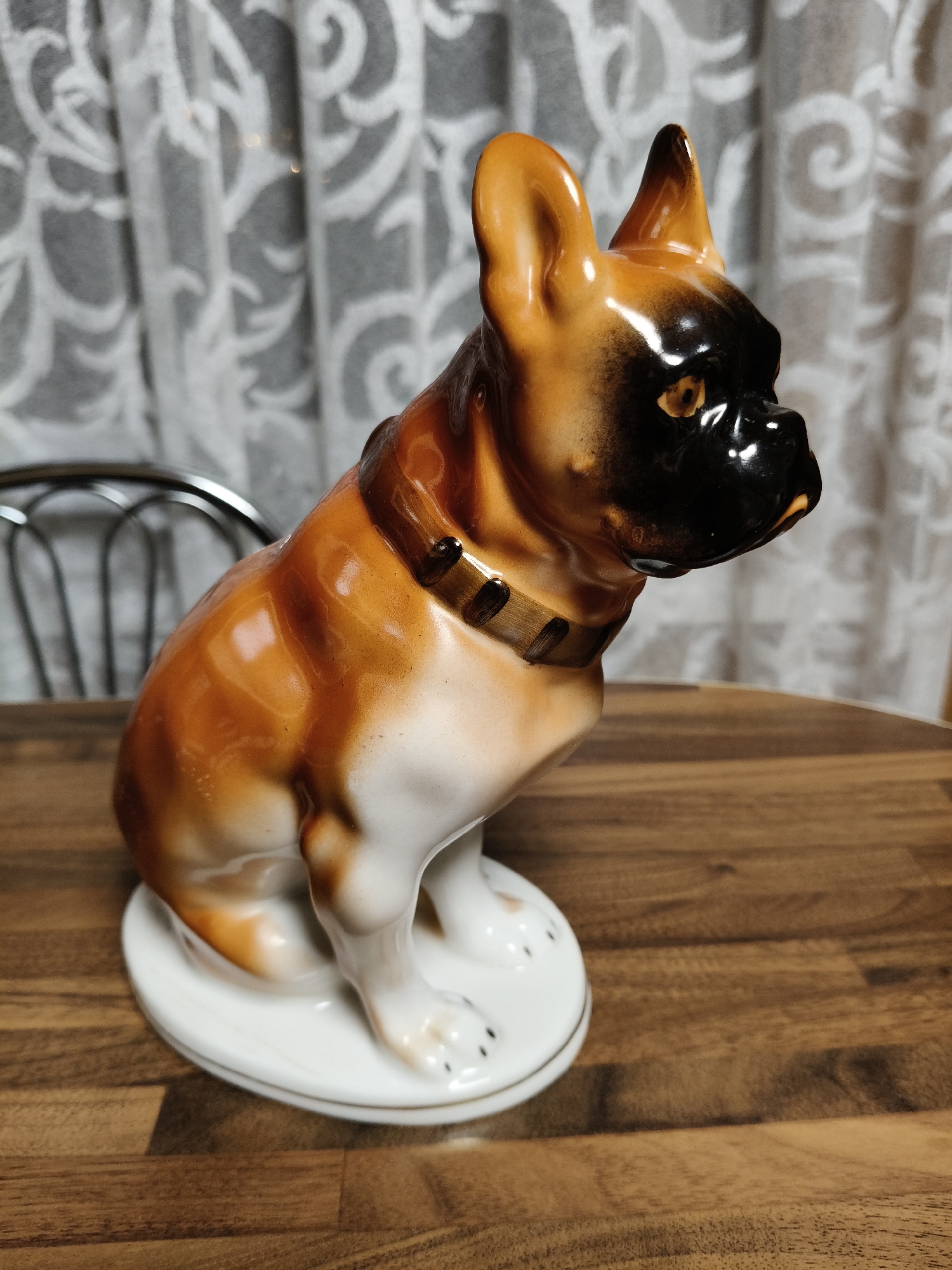 A ceramic figurine of a brindle and fawn-colored dog, resembling a French Bulldog or Boxer, is positioned on a light brown wooden surface. The dog figurine is predominantly tan-brown with darker brown markings, a prominent black muzzle and face mask, and white markings on its chest and paws. It is depicted in a sitting posture, looking towards the right side of the frame, with upright ears and visible eyes. A brown collar with dark brown accents is depicted around its neck. The figurine rests on a white, oval-shaped base with a thin gold rim. The wooden surface displays a distinct horizontal grain pattern. In the background, a translucent white curtain with a faint grey patterned design is visible. To the left, a portion of a metal chair back with a curved, open design is partially visible. The scene is illuminated by soft, ambient light. The location is an interior setting in Unknown, Unknown.