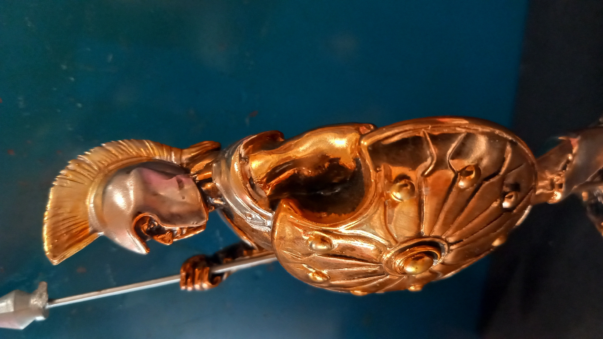 A close-up, low-angle shot shows a metallic figurine of a Spartan warrior, seen in profile from the chest up, with a spear held vertically in its left hand. The figurine is predominantly bronze-colored, with some silver accents on the helmet. The warrior wears a Corinthian helmet with a prominent golden crest, and its chest is protected by a silver cuirass. A round, ornate bronze shield with radiating lines and decorative studs is held in front of its body. The background is a textured, deep teal color with some specks of red and white. The figurine is positioned against a dark, possibly black surface. The overall lighting highlights the metallic sheen of the figurine. There is no discernible location in the city of Kos, Greece.