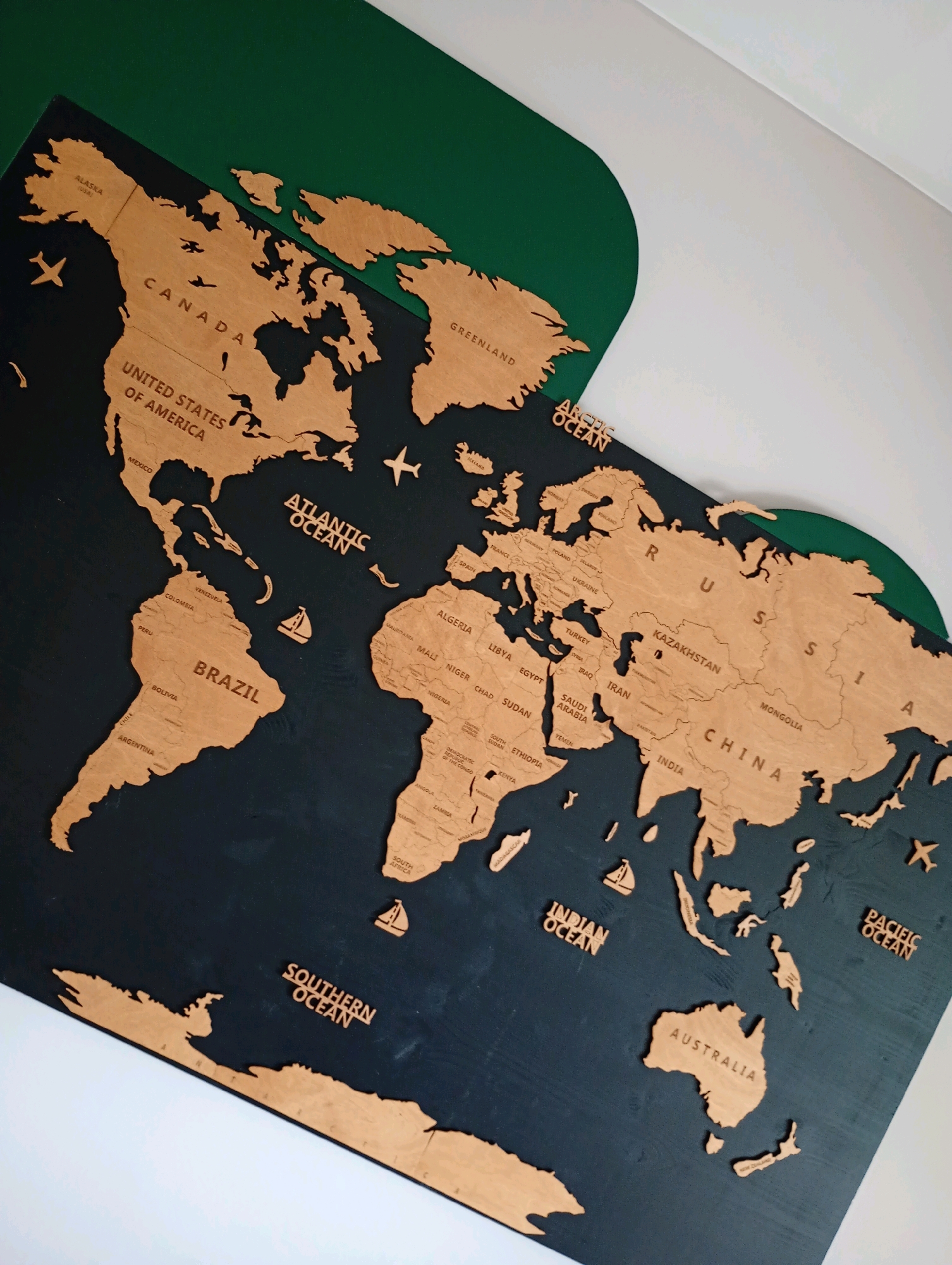 The image displays a wooden world map mounted on a dark background, with sections of green and white material on the upper right. The map features cut-out continents and countries made of light-colored wood, with labels for continents and oceans such as "CANADA", "UNITED STATES OF AMERICA", "BRAZIL", "ATLANTIC OCEAN", "ARCTIC OCEAN", "RUSSIA", "CHINA", "INDIA", "AUSTRALIA", "INDIAN OCEAN", "SOUTHERN OCEAN", and "PACIFIC OCEAN". Small wooden airplane and sailboat figures are placed on the map. The overall scene is consistent with a decorative map possibly found in a residence or public space. The city of Bad Sachsa, Germany, is not depicted or suggested by any element within the image.