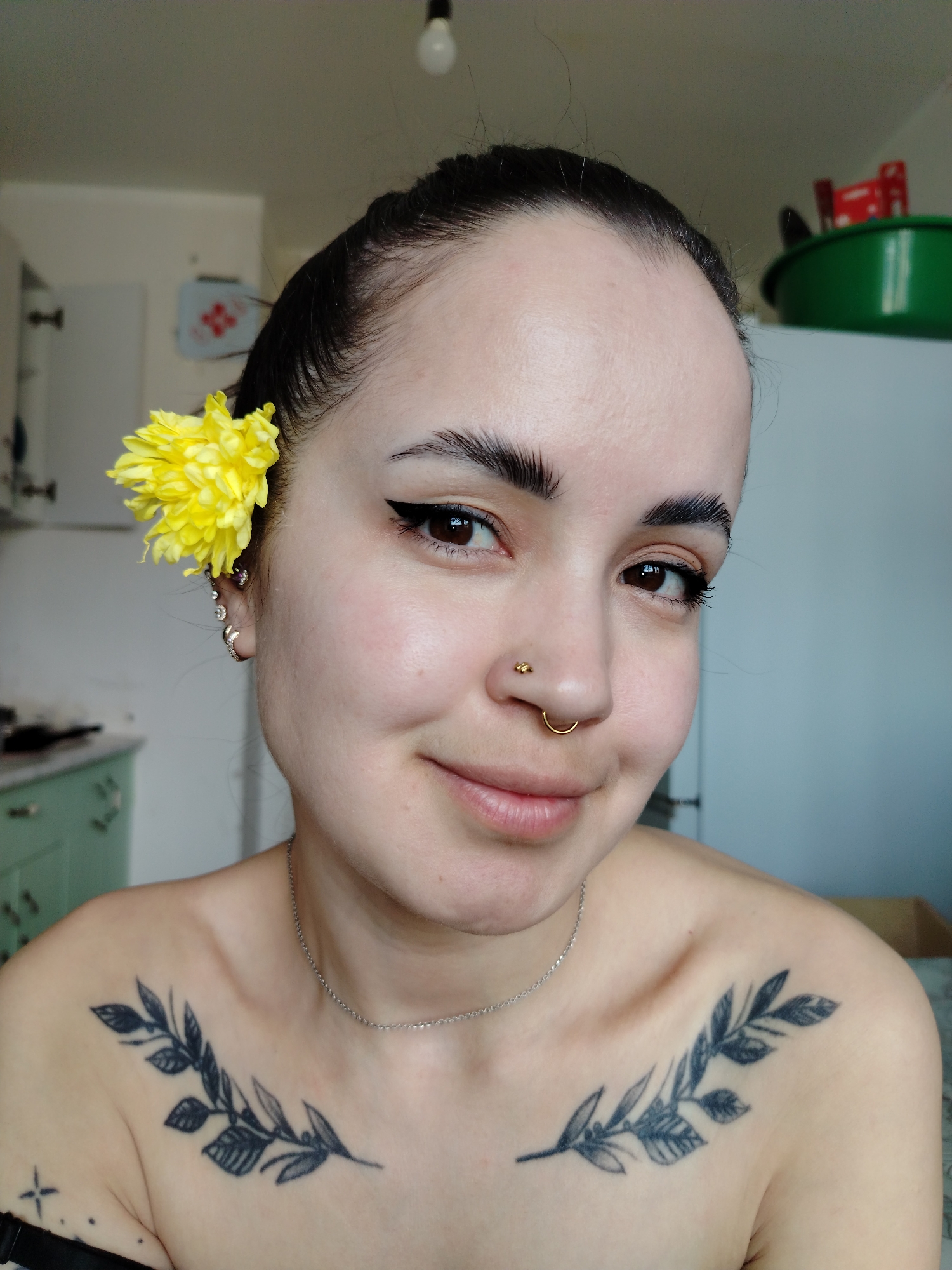 A close-up portrait shows a young woman with dark hair pulled back, adorned with a yellow flower behind her left ear. She has fair skin, well-defined eyebrows, brown eyes with eyeliner, and piercings in her nose and ear. She is wearing a silver necklace. Tattoos of leafy branches decorate her chest.
The background suggests a domestic setting, possibly a kitchen, with cabinets and objects visible.