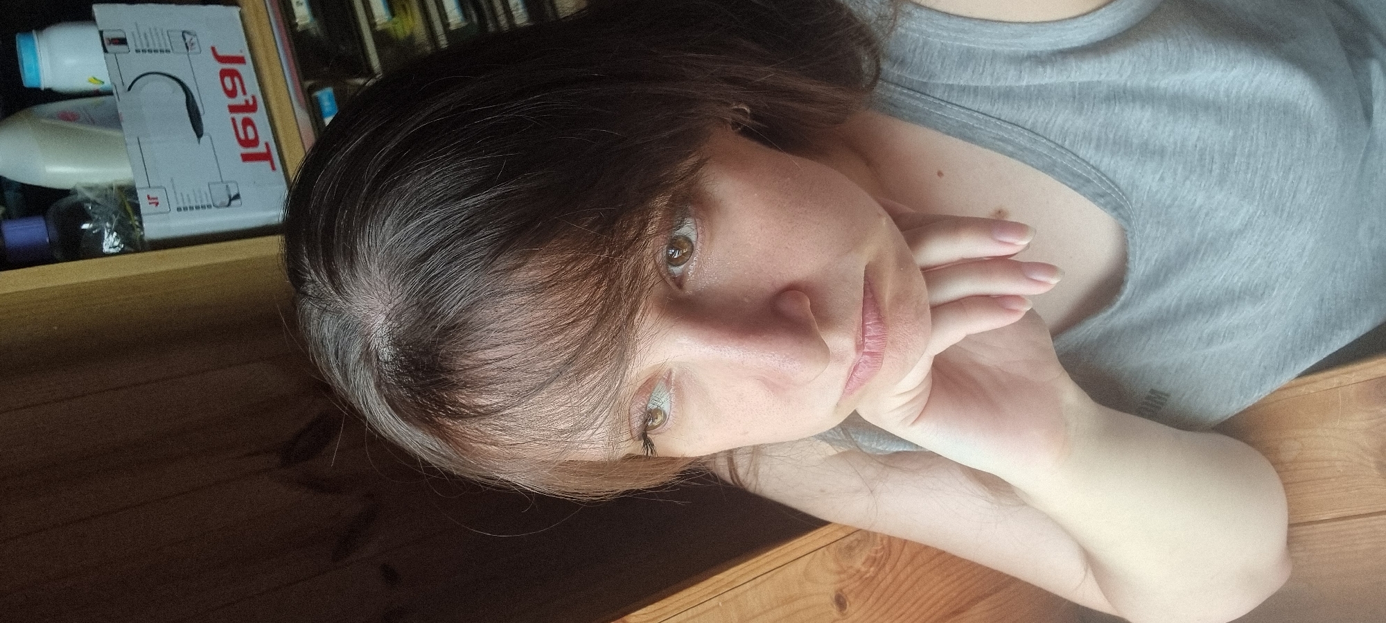 The image shows a woman captured from a slightly elevated perspective, resting on a wooden surface, possibly a floor or low table, in an indoor setting. She has dark brown hair with visible graying at the roots, styled with bangs that frame her face. Her light brown or hazel eyes are open and directed towards the viewer, with a neutral and calm expression. She is wearing a light gray tank top. Her hands are positioned near her face, with fingers gently touching her chin or cheek, suggesting she is supporting her head while lying down.
Strong natural light from the left side of the frame creates noticeable shadows on the wooden surface and highlights her hair and facial features. In the background, behind her head, several objects are visible on a shelf or cabinet. These include various bottles, one with a light blue cap, another white, and a dark purple one. A white cardboard box is also present, displaying reversed text that clearly reads "J619T" and a smaller "Jr." The scene depicts a quiet, relaxed moment in Dunnae-myeon, South Korea.