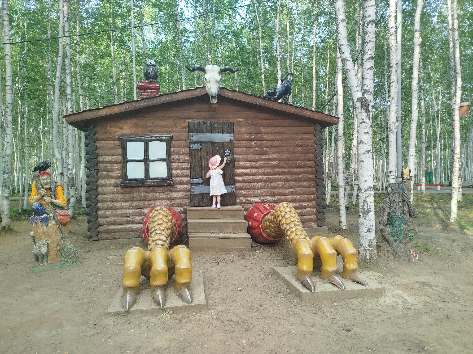 A young girl in a white dress and pink hat stands on the steps of a wooden cabin, reaching for the door knocker. The cabin is decorated with a bull's skull and two owls on its roof, and giant, yellow claws with sharp tips protrude from the ground on either side of the steps. To the left of the cabin, a statue of a witch with a broom and a cat is visible, and to the right, a statue of a tree-like creature with a face stands near the birch trees that surround the area. The ground is dirt and pebbles, and the background is filled with a dense forest of birch trees. This scene appears to be an outdoor attraction, possibly in the city of Mirny, Russia, given the presence of birch trees common to the region.