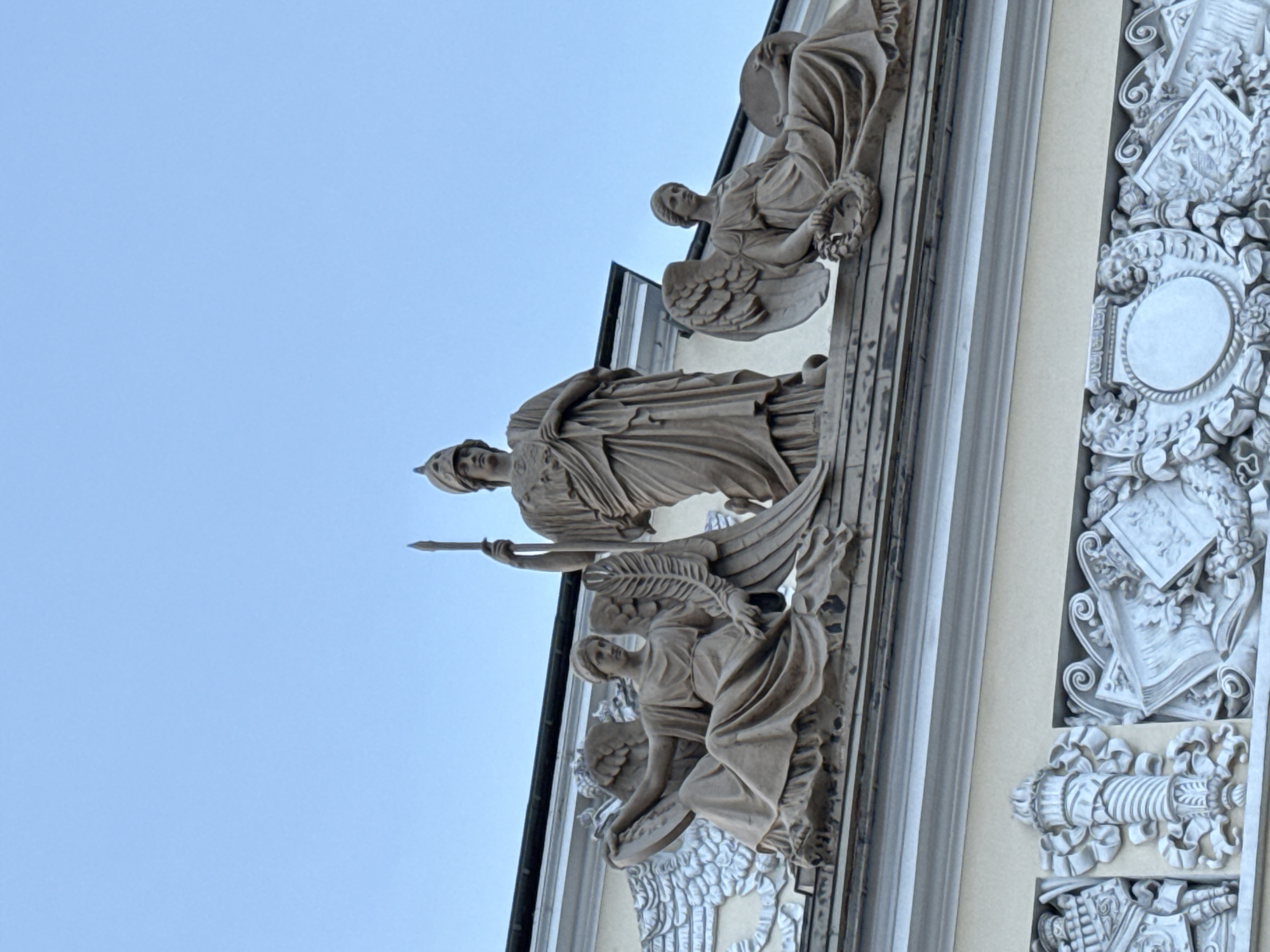 The image shows a fragment of a building facade in Saint Petersburg, Russia, featuring sculpted figures. The main figure is a statue of a woman, likely a deity or allegorical figure, wearing a helmet and holding a spear or scepter. Below her, there's a sculpted angel-like figure with wings. Above the main figure is another sculpted figure, also with wings, holding a wreath or garland. The building is decorated with ornamental details and appears to be made of stone or a similar material. The background is a clear blue sky.