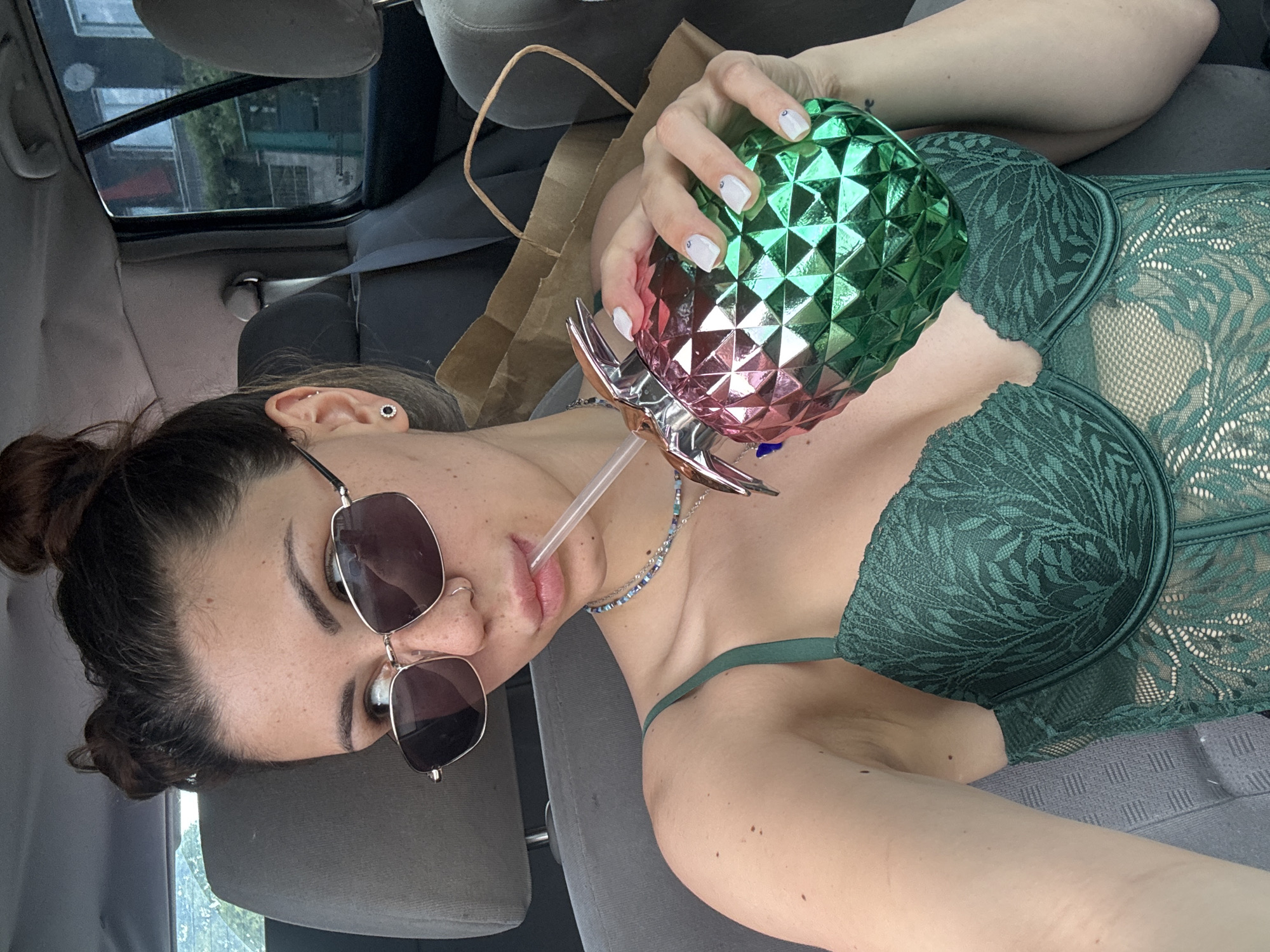 A young woman is depicted lying sideways in what appears to be the passenger seat of a car, viewed from a high angle. She has dark hair pulled back into a bun and is wearing dark sunglasses. Her attire includes a dark green lace bra or bodysuit featuring an intricate leaf-like pattern. She holds a unique pineapple-shaped drink container, which transitions in color from green at the top to pink at the bottom and has a faceted surface. A clear straw extends from the container to her mouth, from which she is drinking.
Her nails are painted white and adorned with small silver embellishments. Multiple piercings are visible on her ear, including a prominent black stud with a silver rim. A delicate beaded necklace in shades of blue and white is draped around her neck.
The car's interior features grey fabric upholstery on the seats and a dark grey sun visor. Through the car window, glimpses of buildings and green foliage are visible, indicating an outdoor, likely urban or suburban, setting during daylight hours. A brown paper bag is partially visible near her arm. The overall atmosphere appears casual and relaxed. No visible text is present in the image. The location context for this media file is Gorna Malina, Bulgaria.