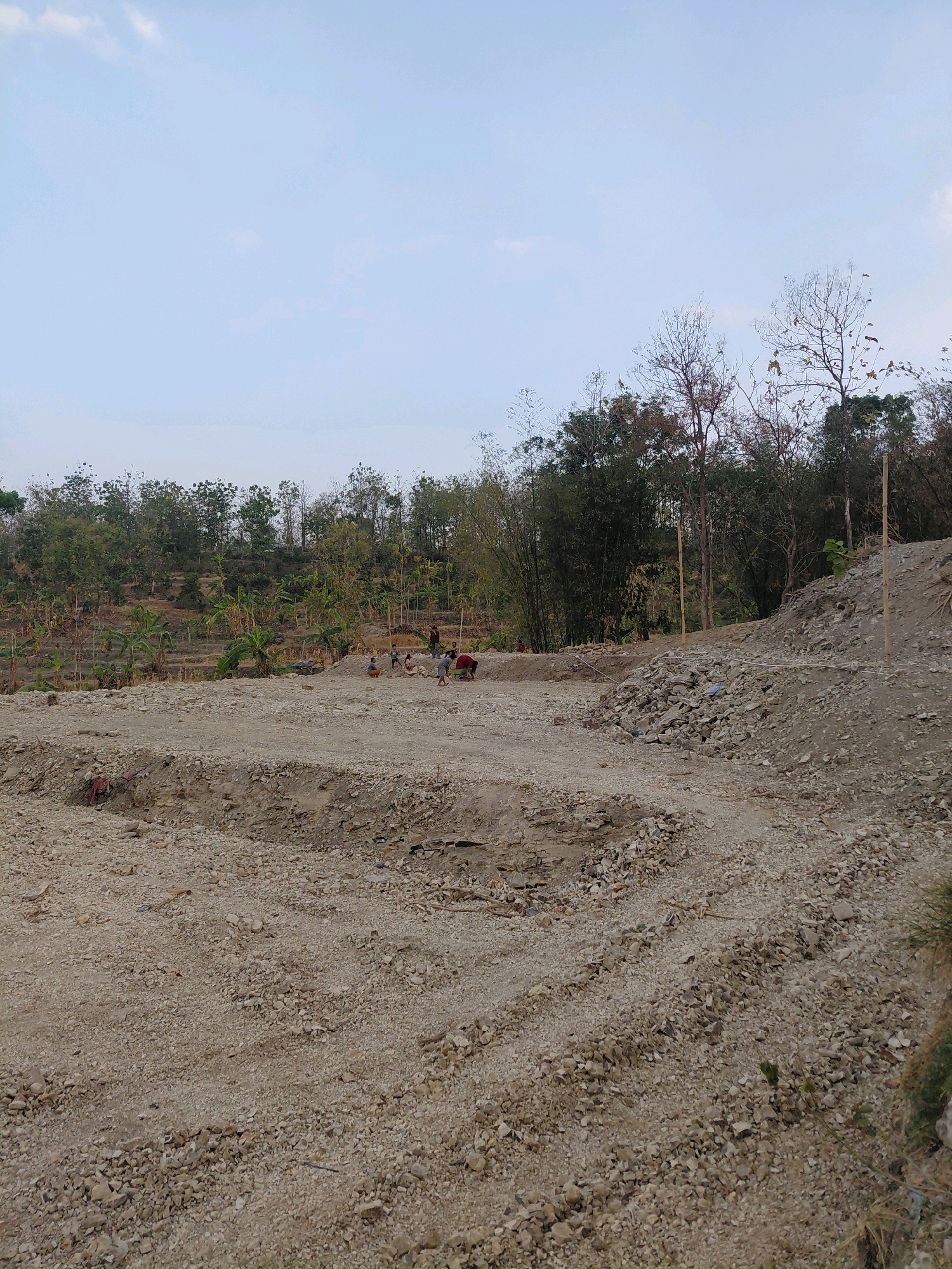 The image depicts an outdoor scene with a clear sky above a landscape that appears to be undergoing some form of construction or excavation. In the foreground, a barren area is covered in gravel and loose soil, with tire tracks visible in the dirt. Further back, a group of people are working in a pit, some of them appearing to be digging or moving materials. Behind them, a terraced hillside is covered with trees and what appear to be agricultural crops, possibly banana trees, with some dry, leafless trees interspersed. The overall impression is one of a rural or semi-rural setting, possibly on the outskirts of a developing area. The image does not provide any specific details that would allow for a definitive location within Bangkok, Indonesia.