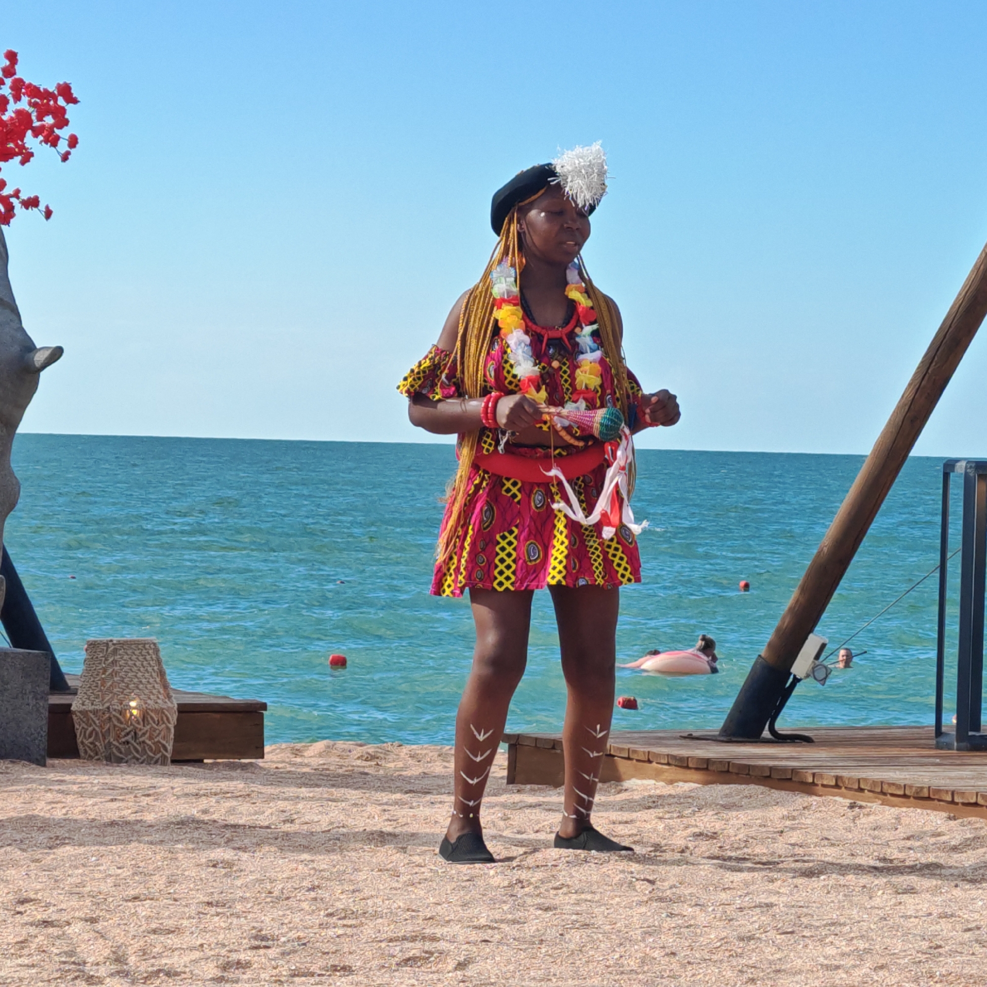 A young Black woman stands on a sandy beach, performing in front of a turquoise ocean under a clear blue sky. She is wearing a vibrant, colorful dress with a geometric pattern in pink, yellow, and black. The dress has off-the-shoulder sleeves and a matching mini skirt. Around her neck, she wears several lei-like necklaces made of colorful artificial flowers and beads, and a red pendant. She also has on red bracelets. A black hat adorns her head, with a white feathery decoration on the side. Her braided blonde hair hangs down her back. She holds a maraca in her right hand and appears to be mid-performance, with her eyes closed and mouth slightly open, suggesting she is singing or chanting. White tribal markings are painted on her lower legs, resembling chevron patterns. She wears simple black slip-on shoes.
To her left, part of a large, gray sculpture resembling an animal's head is visible, with red decorative elements resembling flowers or berries on branches. In the background, a wooden platform extends over the water, with a dark wooden pole supporting an overhead structure. Some red buoys dot the water. Further back, a few people can be seen in the ocean, one on a pink inflatable. A wicker basket with a lamp inside sits on the sand to the left of the wooden platform. The overall scene suggests a daytime event, possibly a cultural festival or performance, taking place in a coastal area. The weather appears to be sunny and warm. The location is identified as Shchyolkine urban-type settlement, Ukraine. There is no visible text in the image.