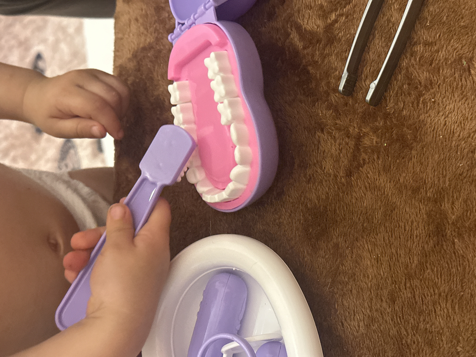 The image shows a child playing with a toy dentist set on a brown surface. The child, whose torso and hands are visible, is holding a purple toothbrush and interacting with the toy mouth, which is also purple with pink gums and white teeth. A white tray containing additional purple dental tools is nearby. To the right of the toy mouth is a pair of metal tweezers. The background is a textured brown surface. The image does not contain the information to provide a location in the city of Kharnavy, Russia.