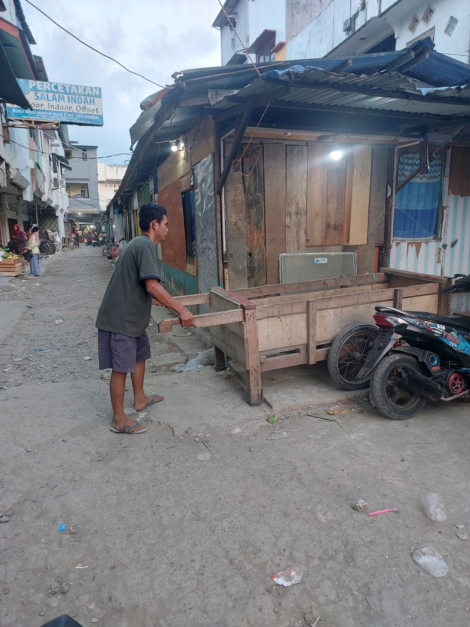 The image depicts a street scene in Ambon, Indonesia, characterized by its unpaved, rough ground, and modest, somewhat dilapidated structures. In the foreground, a man wearing a dark t-shirt and dark shorts, along with flip-flops, is pushing a wooden cart. The cart appears to be a simple, unpainted wooden structure with two large wheels, possibly used for transporting goods. Beside the cart, a dark-colored motorcycle is parked.
The background reveals a narrow street lined with buildings. On the left side, a sign is visible that reads "PERCETAKAN SALAM INDAH" followed by "oor, Indoor, Offset". Below this, smaller text indicates "RUKO BAYU MERAH BLOK R NO. 151 AMBON". This sign suggests the presence of a printing business in the vicinity. Further down the street, other people and market stalls are discernible, indicating a commercial or residential area. The buildings themselves appear to be constructed from various materials, including wood and corrugated metal, and some show signs of age and wear. The overall environment suggests a bustling, everyday setting in a part of the city that may be older or less developed. The lighting suggests it is daytime, possibly overcast.
