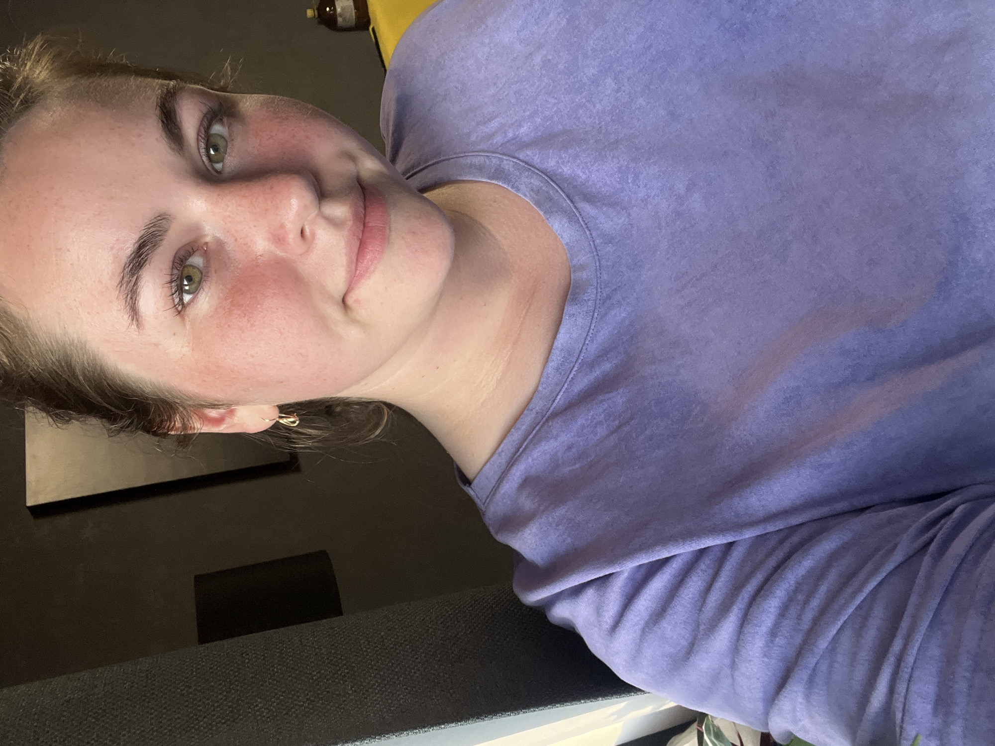 A young woman with fair skin and freckles is pictured from the chest up, looking directly at the camera with a slight smile. She has light brown hair pulled back, green eyes, and her cheeks are flushed pink. She is wearing a light purple t-shirt. In the background, to the left, is a dark gray textured surface, possibly a curtain or upholstered furniture. To the right, a dark wall is visible, with a rectangular shape casting a shadow. A small, shiny gold hoop earring is visible in her left ear.