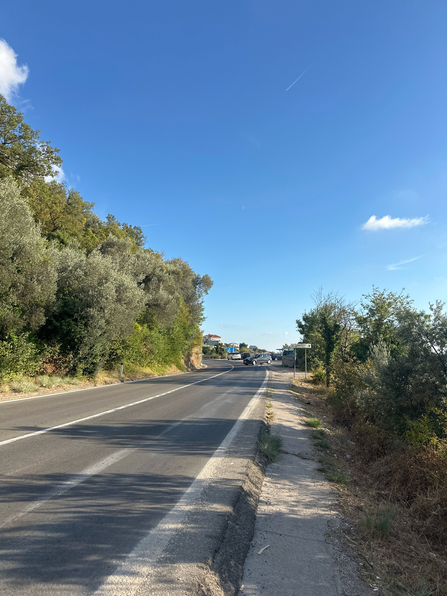 The image shows a paved road curving to the left, bordered by a dense growth of trees and vegetation on the left and a concrete sidewalk and more vegetation on the right. The road has clear white lane markings. Several cars are visible on the road in the distance, with one parked on the right shoulder. A signpost with an arrow pointing to the right is located on the right side of the road. The sky is bright blue with a few wispy clouds and a contrail from an airplane. The scene appears to be a scenic route, possibly in a Mediterranean or coastal area. The location is identified as the city of Drobnići, Montenegro.