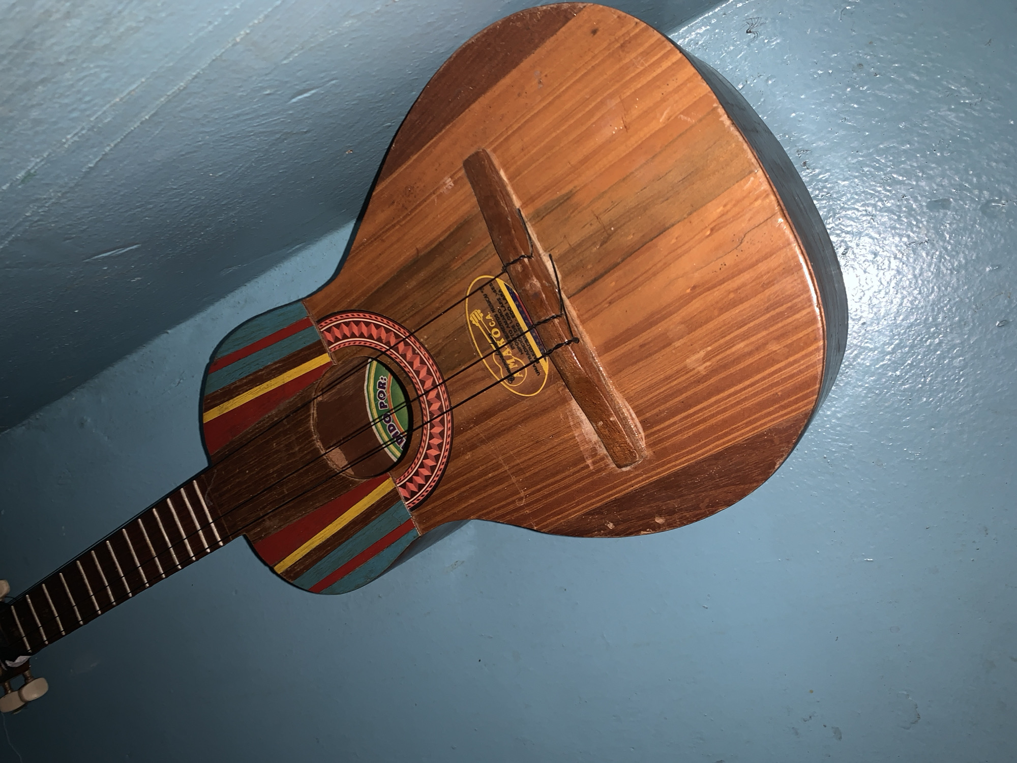 A small, handcrafted wooden string instrument, possibly a Venezuelan cuatro, is prominently featured against a textured, light blue background, likely a wall or ceiling. The instrument's body is made of light brown wood, exhibiting visible grain and some wear. Its soundhole is encircled by a vibrant, multi-colored rosette featuring a geometric pattern in red, yellow, blue, black, and white. Similar colorful stripes adorn the lower edge of the instrument's body. Four strings are visible, secured to a simple wooden bridge.
An oval-shaped yellow label is affixed to the instrument's body, bearing partially visible text. The OCR identifies phrases such as "AMAROCA," "YOS MUSICAI ENTOS" (likely "INSTRUMENTOS MUSICALES"), "UNA TRADICIÓ D" (likely "UNA TRADICIÓN DE"), and "TO EDO LARA" (referring to Estado Lara). Other visible text includes "IDO POR" around the soundhole and "CION DE LOS LARSSES" on the label. The background's consistent texture and color suggest an indoor setting, possibly in Santa Ana, Venezuela, where such folk instruments are common. The lighting produces some reflections on the blue surface.