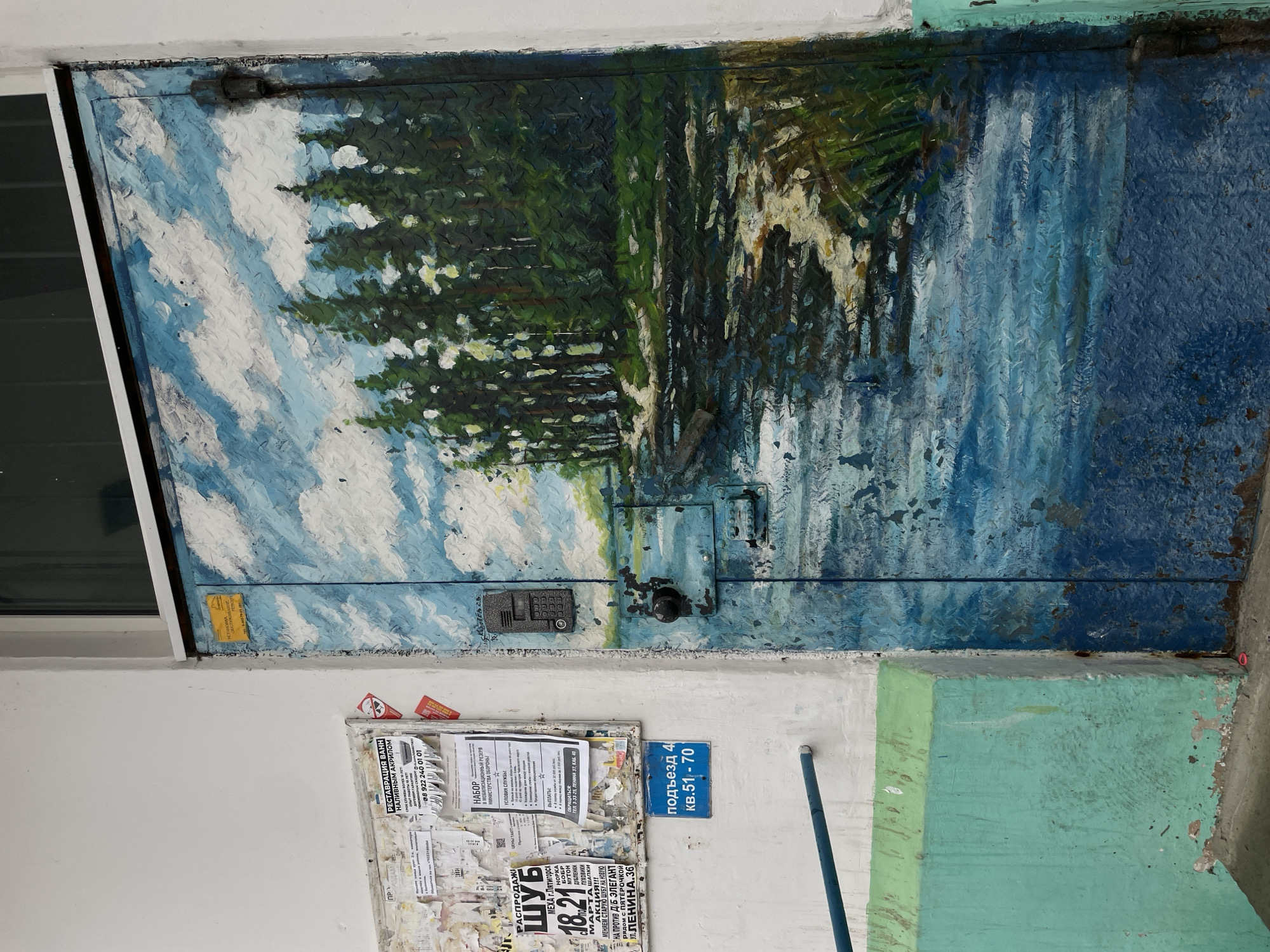 An exterior shot depicts the entrance to "ПОДЪЕЗД 4 кв.51-70" (Entrance 4 apt. 51-70) of an apartment building in Chaikovsky, Russia. The scene is dominated by a heavy metal entrance door, painted with a landscape featuring a blue sky with white clouds at the top, a dense forest of dark green trees in the middle, and a body of water reflecting the sky and trees in the foreground, with some green foliage along its edge. The door's textured surface is visible through the paint, indicating wear. A black digital intercom keypad is mounted on the upper left of the door, and below it is a black cylindrical door handle with a keyhole. Prominent hinges are visible on the door's right side.
To the left of the door, a white painted wall hosts an irregularly shaped bulletin board covered with numerous overlapping papers and advertisements in Cyrillic. These include notices for a "РАСПРОДАЖИ ШУБ" (Fur Sale) at "УЛ.ЛЕНИНА,36" (Lenin Street, 36) and a "НАБОР В МОБИЛИЗАЦИОННЫЙ РЕЗЕРВ МИНИСТЕРСТВА ОБОРОНЫ" (Recruitment to the Mobilization Reserve of the Ministry of Defense) explicitly mentioning "г. Чайковский" (Chaikovsky city). Below the bulletin board, a blue rectangular sign with white text identifies the entrance as "ПОДЪЕЗД 4 кв.51-70". A horizontal blue metal railing is affixed to the wall below this sign. The lower portion of the wall is painted a light turquoise/mint green, showing significant wear, peeling paint, and dirt accumulation. Above the door and wall, a dark window with a white frame is visible. The ground surface, partially visible at the bottom, appears to be concrete or asphalt, with a small pink object resting near the door.