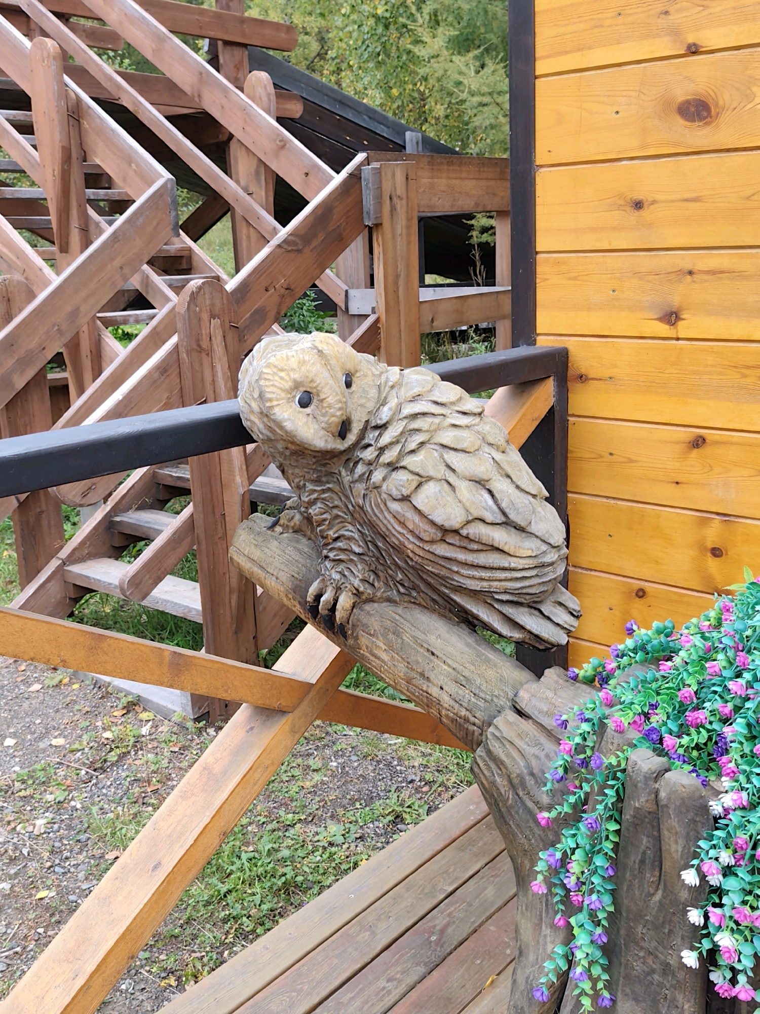 A carved wooden owl sculpture is perched on a log, which is part of a decorative element on the exterior of a wooden building in Listvyanka, Russia. The building's exterior is clad in horizontal wooden planks with a yellow-orange stain. To the left of the sculpture is a rustic wooden staircase leading upwards. In the foreground and to the right of the owl, artificial flowering vines with purple, pink, and white blossoms cascade down a wooden post. The ground is covered with gravel and scattered leaves. The overall impression is of a decorative outdoor scene at a rustic establishment.