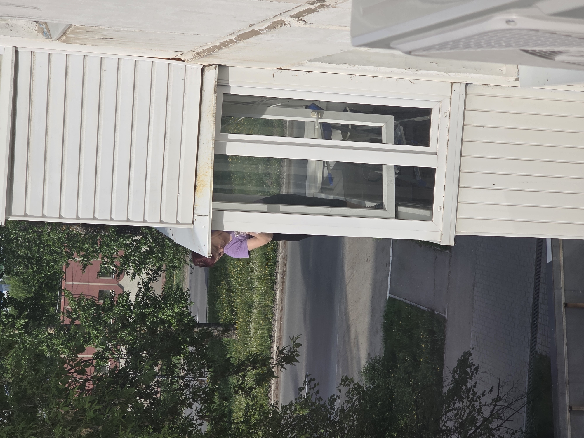 The image depicts an exterior view from a building window in Chaikovsky, Russia. A person with dark hair and wearing a purple shirt is leaning out the window. The window frame is white and has a double-hung design. The exterior of the building is white with a corrugated metal facade. A street runs parallel to the building, with grass and trees lining the side of the road. In the distance, a red brick building is visible through the trees. The overall impression is that the photo was taken on a sunny day.
