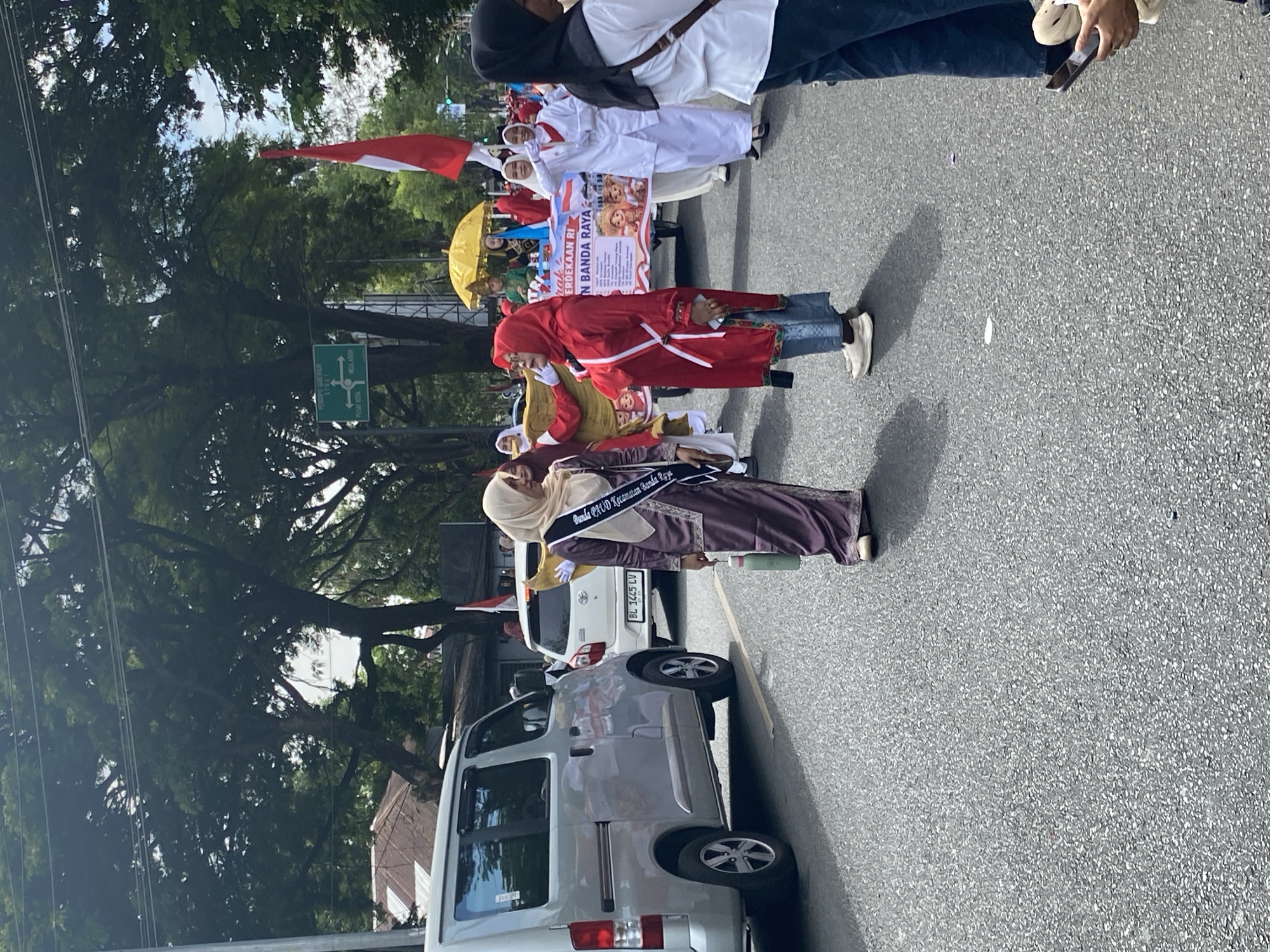The image depicts a street parade in Banda Aceh, Indonesia. In the foreground, two women are walking on the asphalt road. The woman on the left is wearing a long, purple dress and a beige hijab, with a sash across her chest that reads "JALAN JALAN KECAMATAN BANDA RAYA". She is holding a light green water bottle in her right hand. The woman on the right is wearing a red hijab and a red outfit with a white sash. She is wearing blue jeans and white sneakers. Behind them, several other people are visible, some in white uniforms and headscarves, suggesting they are part of a procession or performance. A white van is parked on the left side of the road, and a white car is seen in the background. Further down the street, a yellow umbrella is being held aloft, and a red and white Indonesian flag is displayed. Green trees line the street, and a street sign is visible in the upper right corner, indicating directions within the city. The atmosphere appears to be festive, likely a public holiday or celebration.