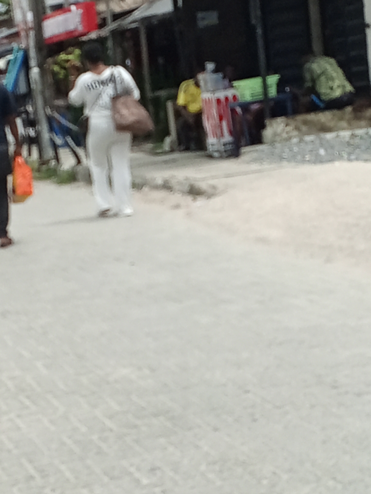 The image shows a street scene, possibly in a city, with several people and objects. A person in a white outfit walks away from the camera on the left, carrying a brown bag. Beside them is another person. In the background, figures are seated, and there are various objects like plastic containers, a white container, a red box, and what appears to be street furniture. The setting is an outdoor environment. The location is Unknown, Unknown.
