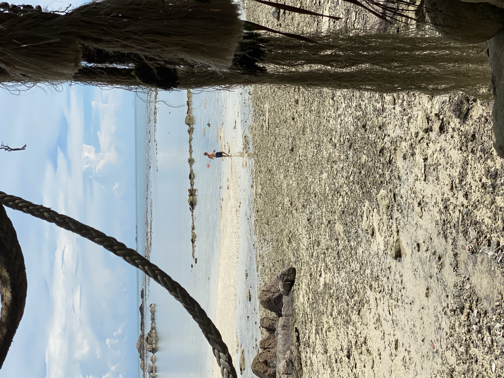 A boy is fishing with a red net in a shallow body of water near the shore at low tide. The beach is composed of sand, small rocks, and debris. In the foreground, thick ropes hang down, obscuring part of the view. In the background, the ocean extends to the horizon, with a cloudy sky above. The location is identified as Ko Pha Ngan, Thailand.