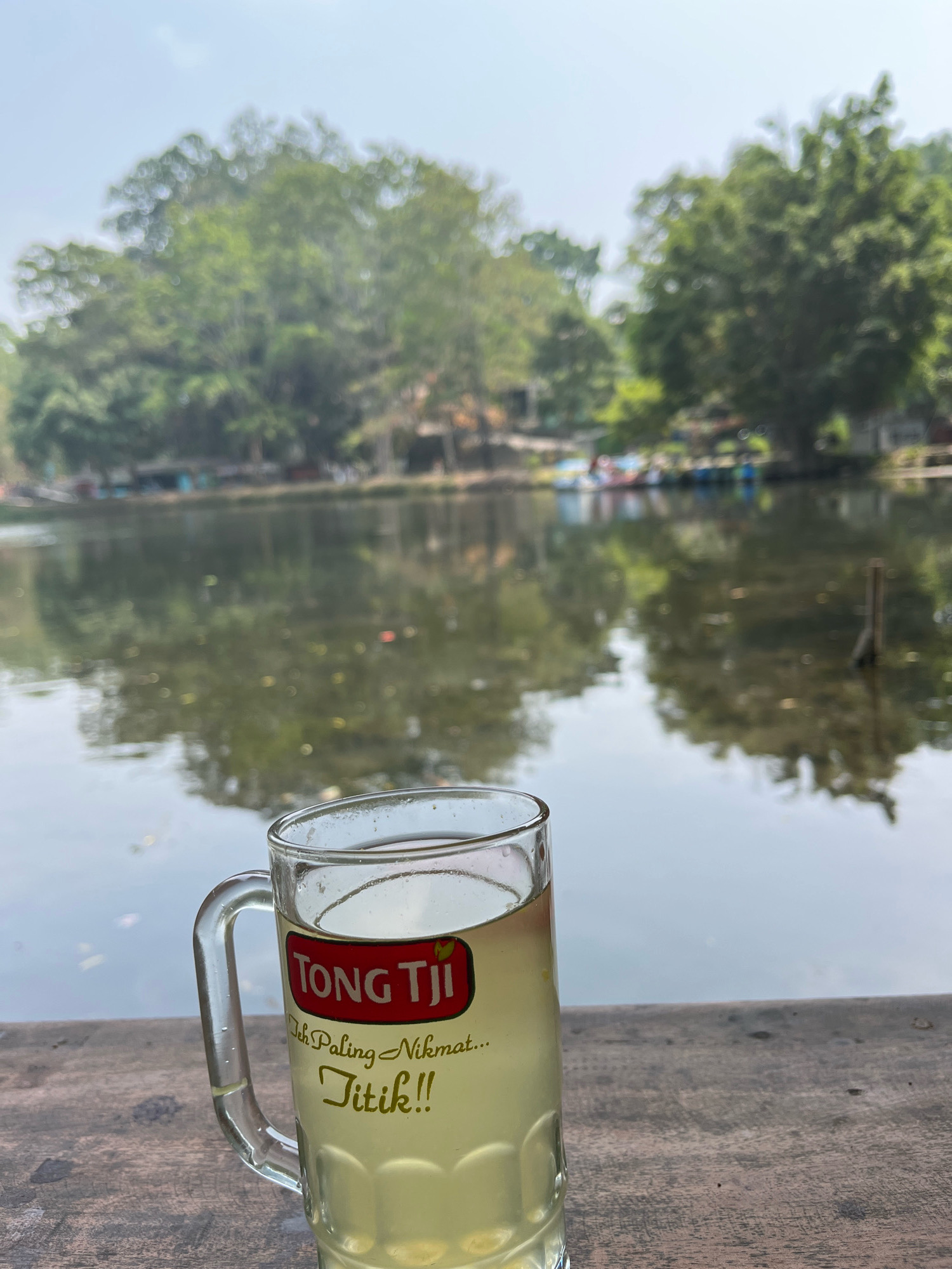 A glass of Tong Tji tea sits on a wooden surface in front of a body of water. The tea is a pale yellow color and the glass has a logo that reads "TONG TJI Teh Paling Nikmat... Titik!!". The background shows trees and a pale sky, with reflections of the trees on the water. The setting appears to be outdoors, possibly in Tegalwaton, Indonesia, a place known for its natural scenery and local tea brands.
