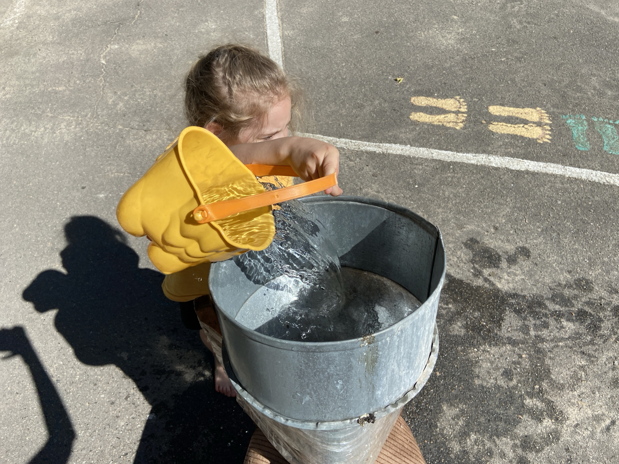 The image shows a young child pouring water from a yellow bucket into a large metal container. The child, with blond hair pulled back, is wearing a yellow shirt. Their right hand holds the handle of the yellow bucket, tilted to let water stream out. The metal container appears to be a galvanized steel tub, possibly an old wash tub or a large planter. The tub is placed on a surface that suggests an outdoor location, with asphalt or concrete visible in the background. Faint chalk markings, resembling footprints in yellow and turquoise, are visible on the ground, suggesting this might be a playground or a recreational area. The lighting indicates it's daytime, with strong sunlight casting shadows, including the silhouette of the child and their arms. The overall mood is one of play and simple activity, likely on a warm day. There is no visible text in the image.